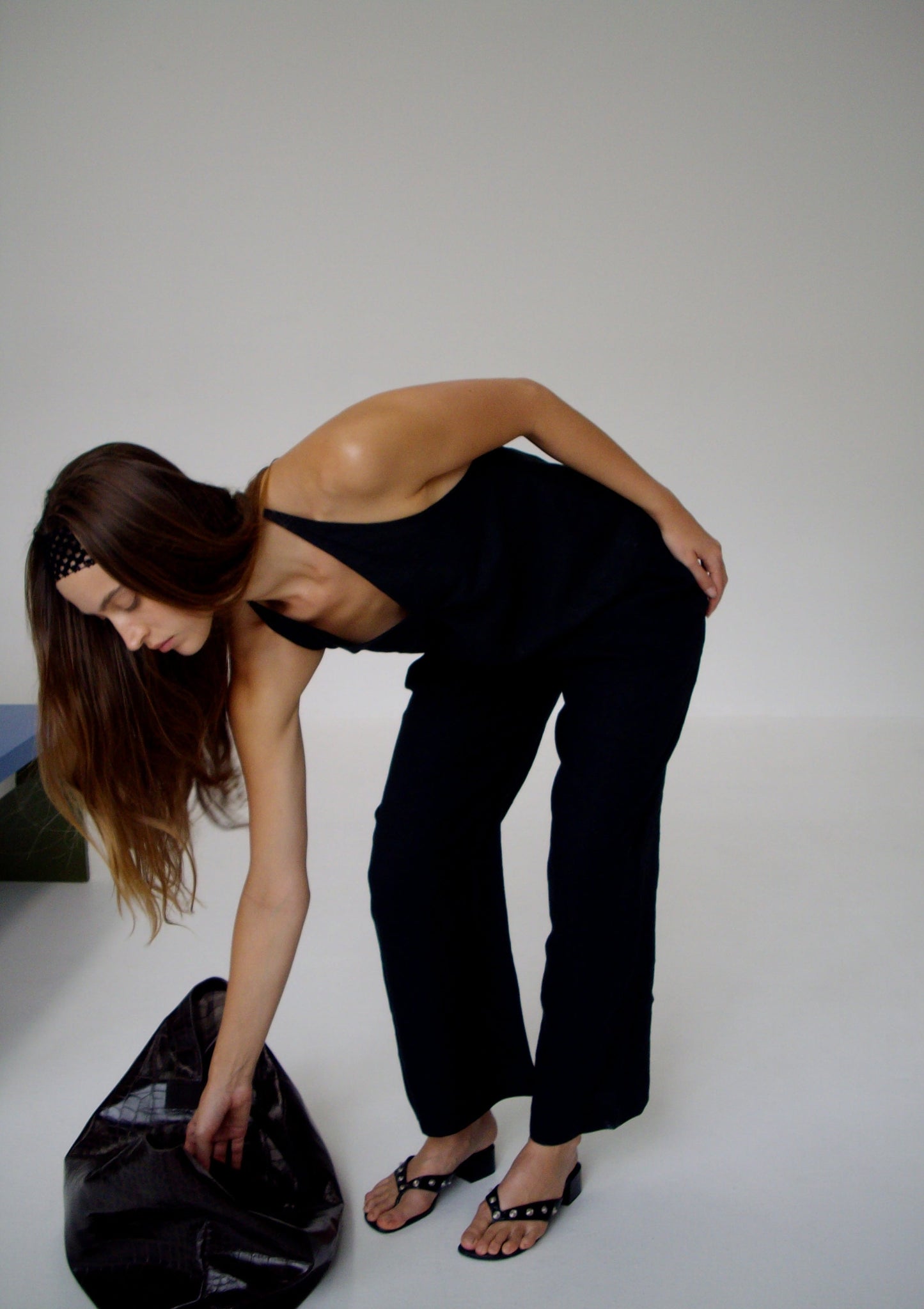 A woman with long brown hair bends down to pick up a large black bag in a minimal, light room. She wears the Linen Ease Trouser - Black with a black sleeveless top and simple sandals.