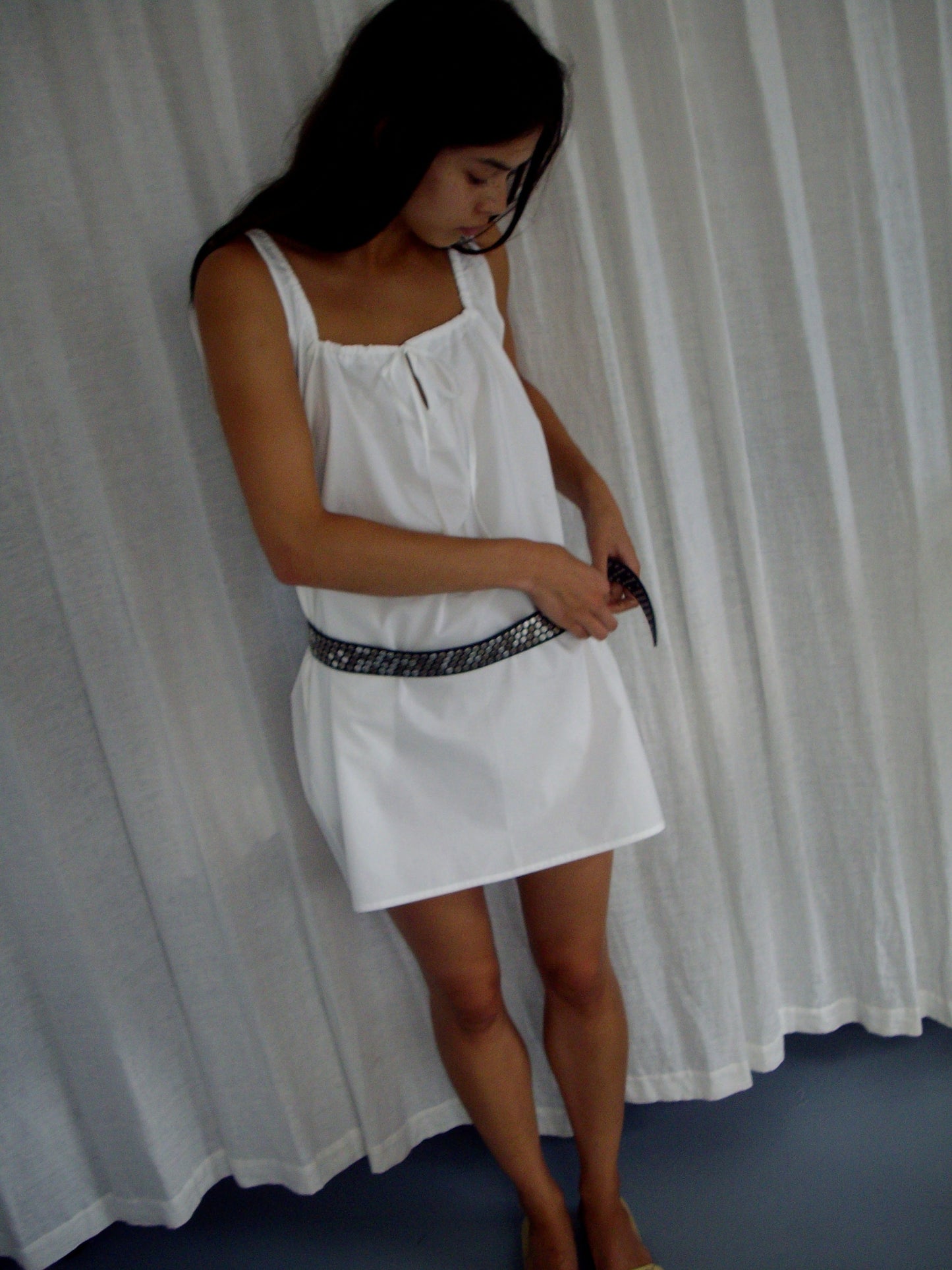 A woman stands barefoot indoors, fastening a black belt around her waist while wearing The Bow Dress - White by Deiji Studios. A white curtain hangs in the background.
