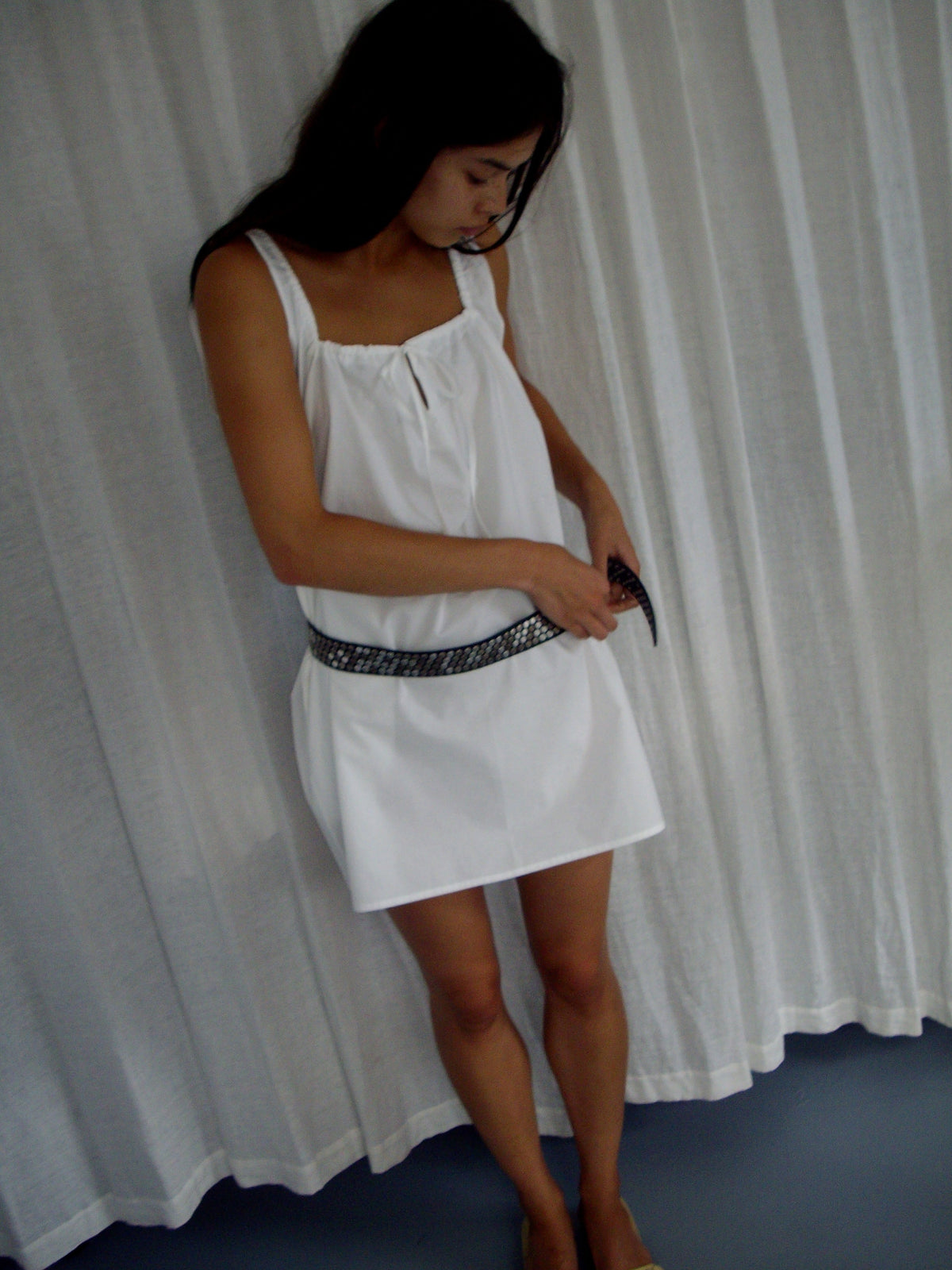 A woman stands barefoot indoors, fastening a black belt around her waist while wearing The Bow Dress - White by Deiji Studios. A white curtain hangs in the background.