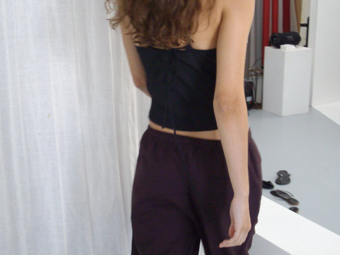 A person with wavy brown hair, wearing a black top and the Drawcord Culotte - Burgundy Brown Stripe by Deiji Studios, stands indoors facing away from the camera, surrounded by white curtains, a white platform, and scattered objects on the floor.