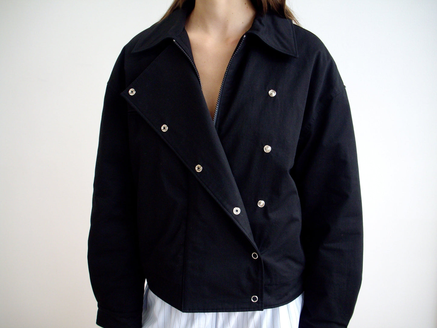 A person wears the Lapel Jacket - Black, featuring an asymmetrical lapel, silver buttons, and wide collar, paired with a light pleated skirt. The style echoes Deiji Studios' minimalist designs against a plain white background.
