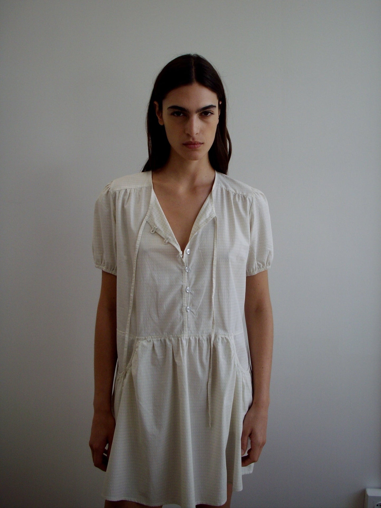 A woman with long dark hair stands against a white wall, wearing the Pocket Mini Dress—a light short-sleeved, button-front dress with a loose, relaxed fit. She gazes at the camera with a neutral expression.