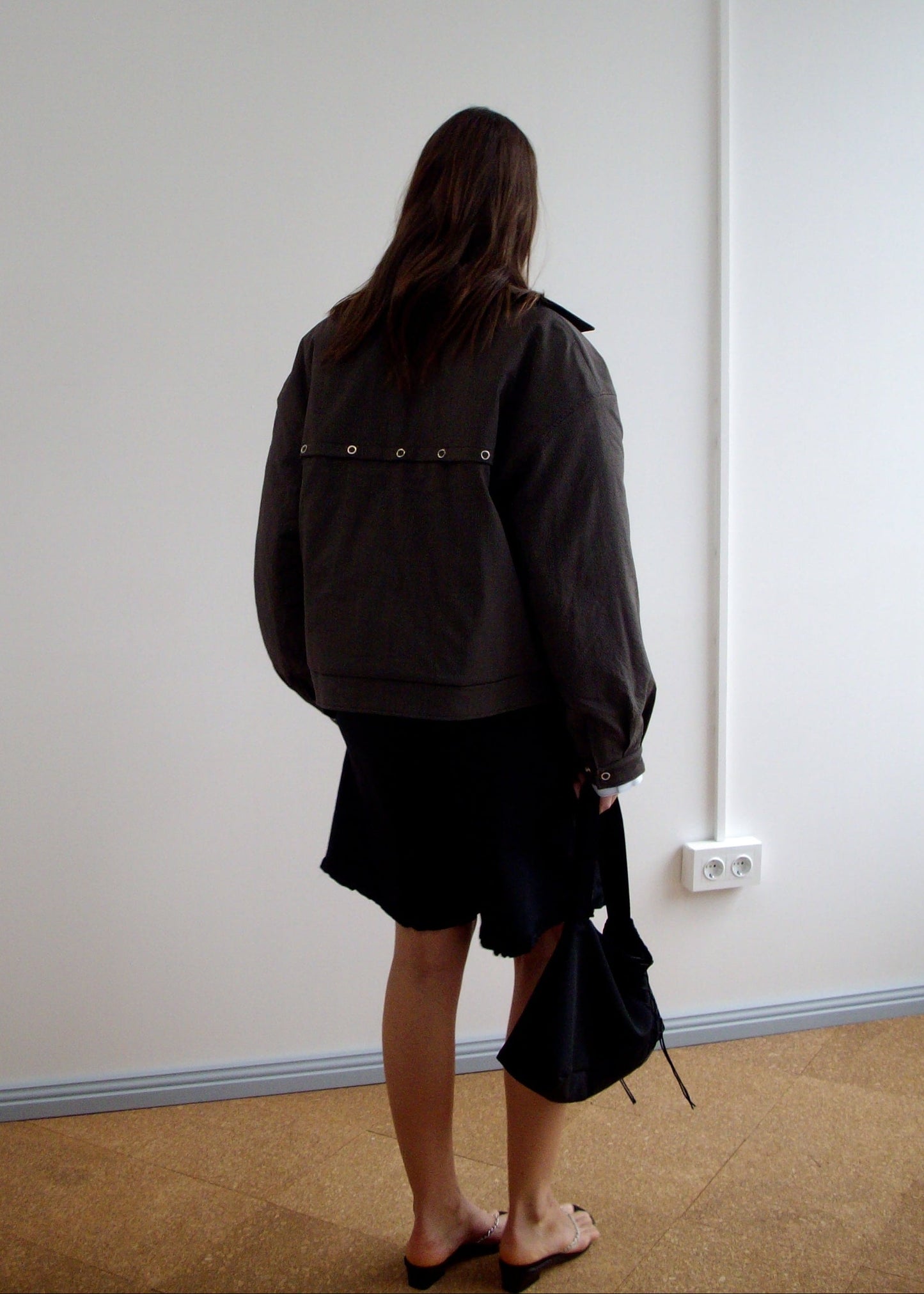 A person with long hair faces away, wearing a dark oversized jacket, the Flounce Short - Black with an elastic waist, and holding a black bag. Indoors near a white wall with outlets, they wear black slide sandals.