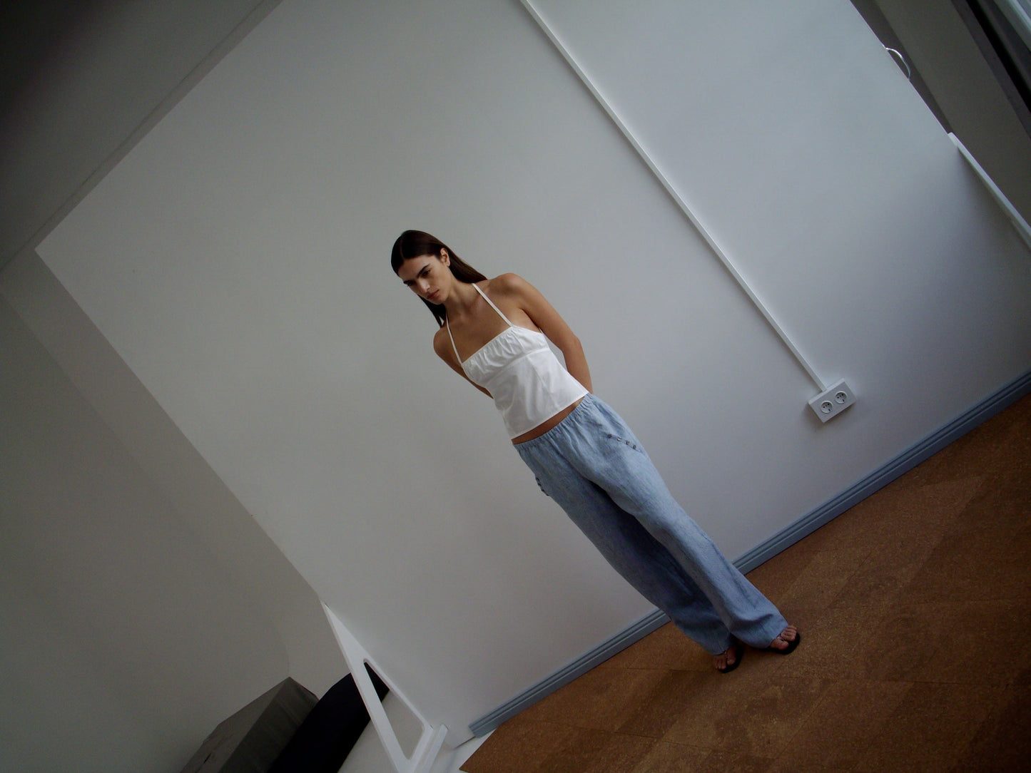A woman stands indoors against a plain white wall, wearing the Strap Top - White and loose light blue pants. She looks down with her hands behind her back; the room features minimal furniture and soft lighting.