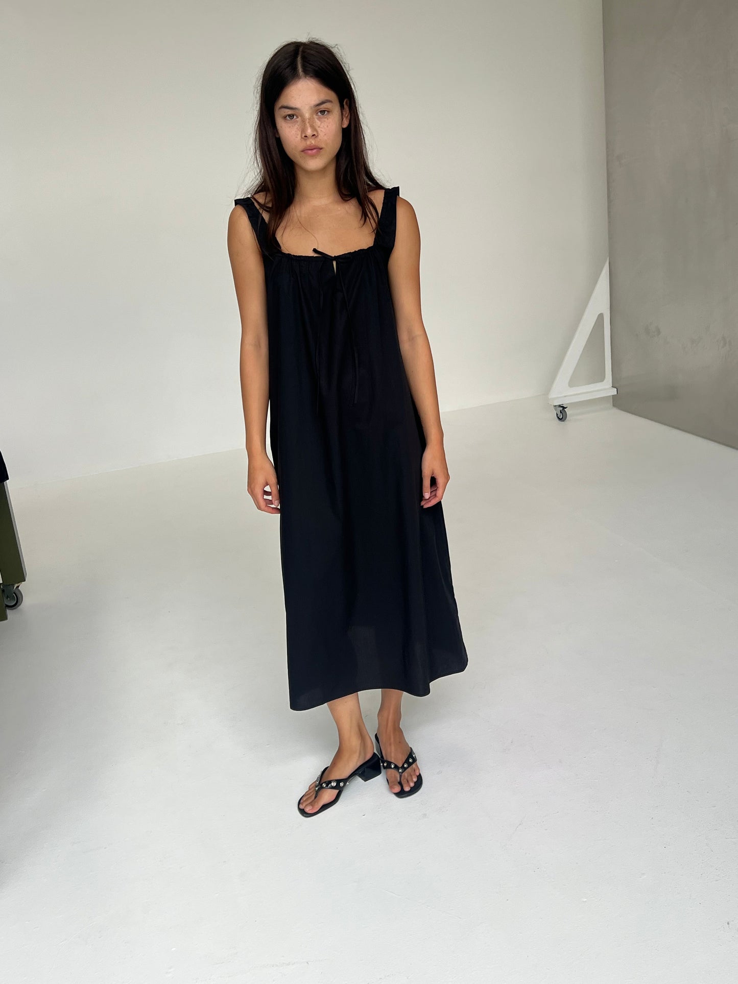 A woman with long dark hair stands in a minimal white room, wearing The Paper Dress - Black by Deiji Studios and black sandals. She looks forward with a neutral expression, her arms relaxed at her sides.