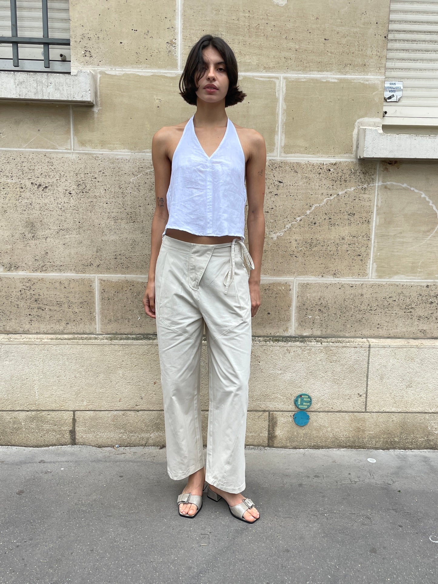 A person stands by a beige stone wall, wearing a white sleeveless halter-top, Flag Tie Pant - Off White, and sandals. With short dark hair and arms at their sides, they appear relaxed.