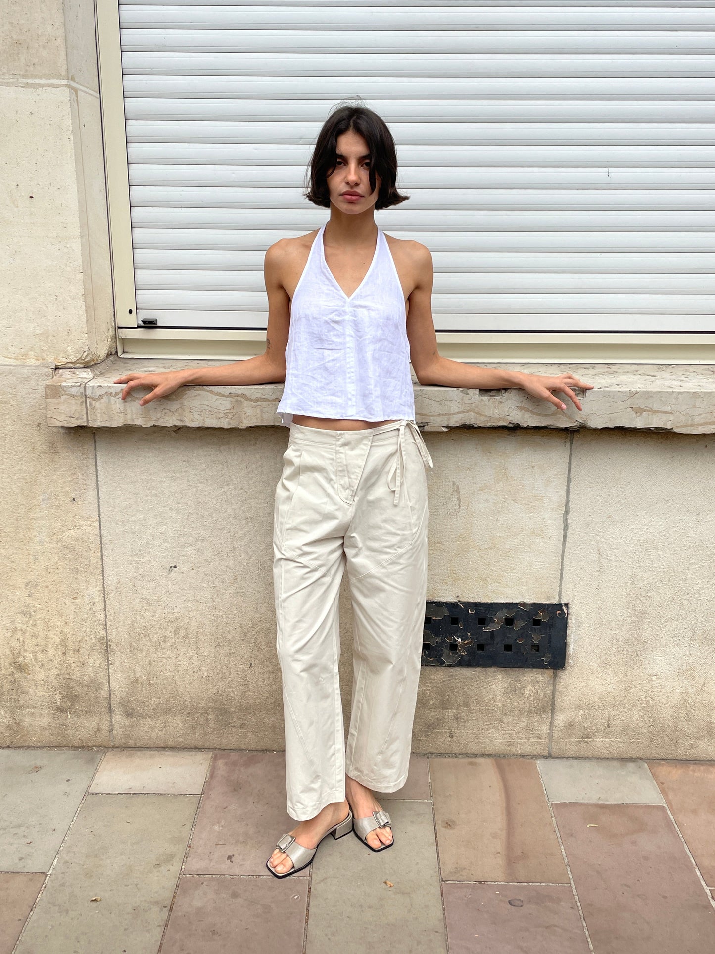 A woman with short dark hair stands against a beige stone wall and closed white shutter. She wears the Linen Pinched Halter Top - White by Deiji Studios, cream wide-leg pants, and sandals, her arms resting on the ledge behind her.