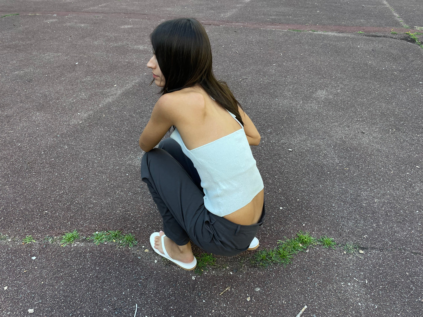 A young woman with long dark hair crouches on outdoor asphalt, seen from behind. She wears a white crop top, the Zip Pant - Charcoal by Deiji Studios, and white flip-flops, gazing to the side.