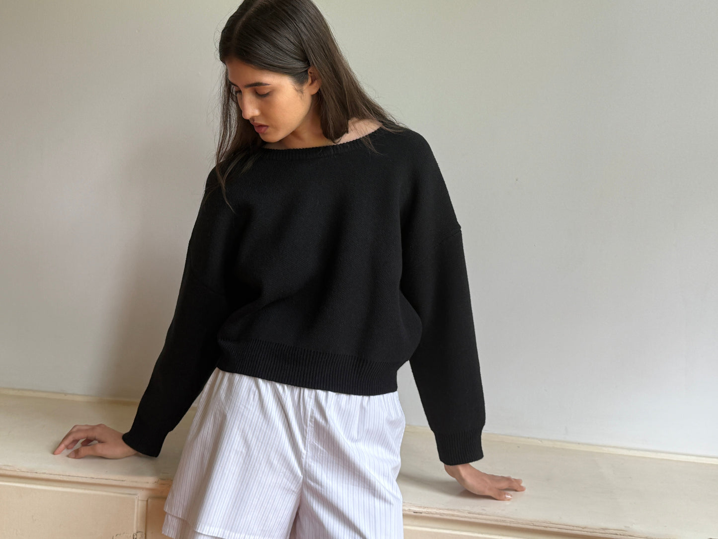A young woman with long dark hair wears the 0 Pullover - Black, paired with white shorts, standing indoors against a plain light wall and looking down to her left with one hand resting on a surface.
