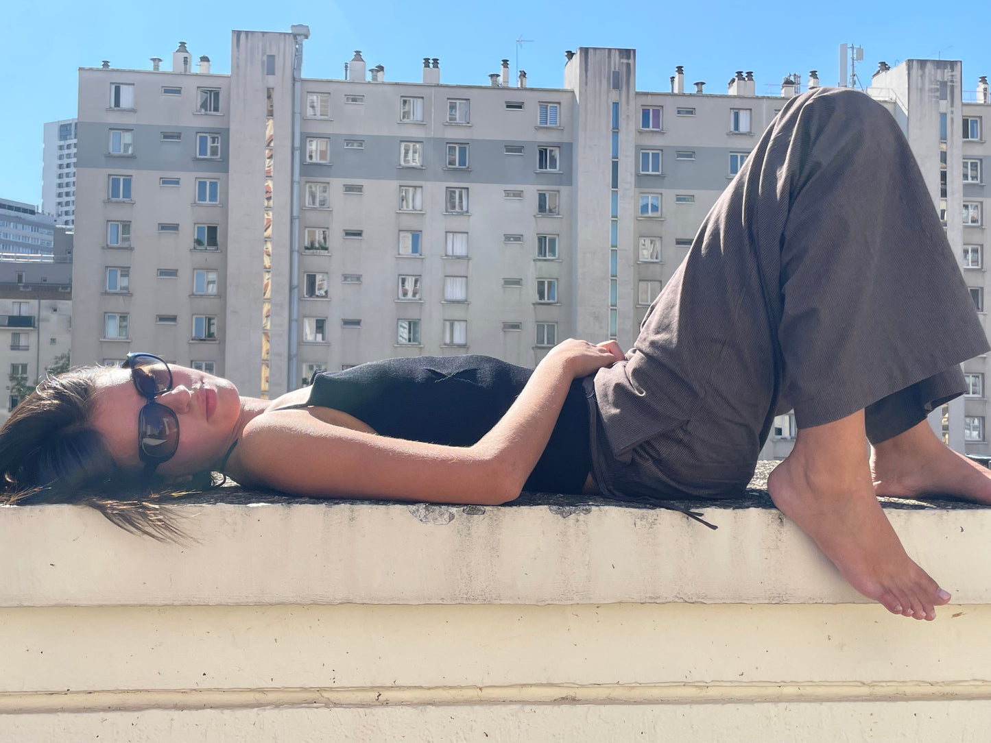 A woman in sunglasses and a Fine Knot Tank - Black with gray pants lies barefoot on a ledge, knees bent. Behind her, apartment buildings rise under a clear blue sky.
