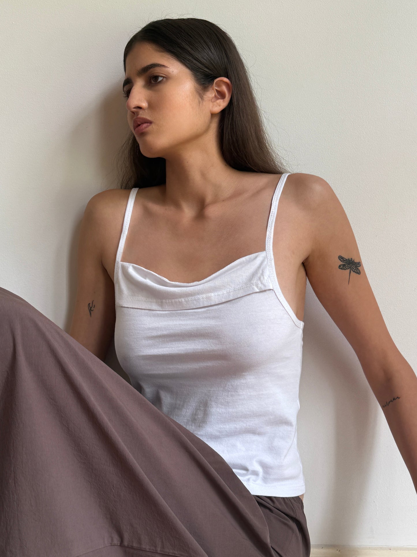 A woman with long dark hair and arm tattoos wears the Simple Tank - White by 0 with brown pants, sitting against a plain white wall and gazing to the side with a neutral expression.