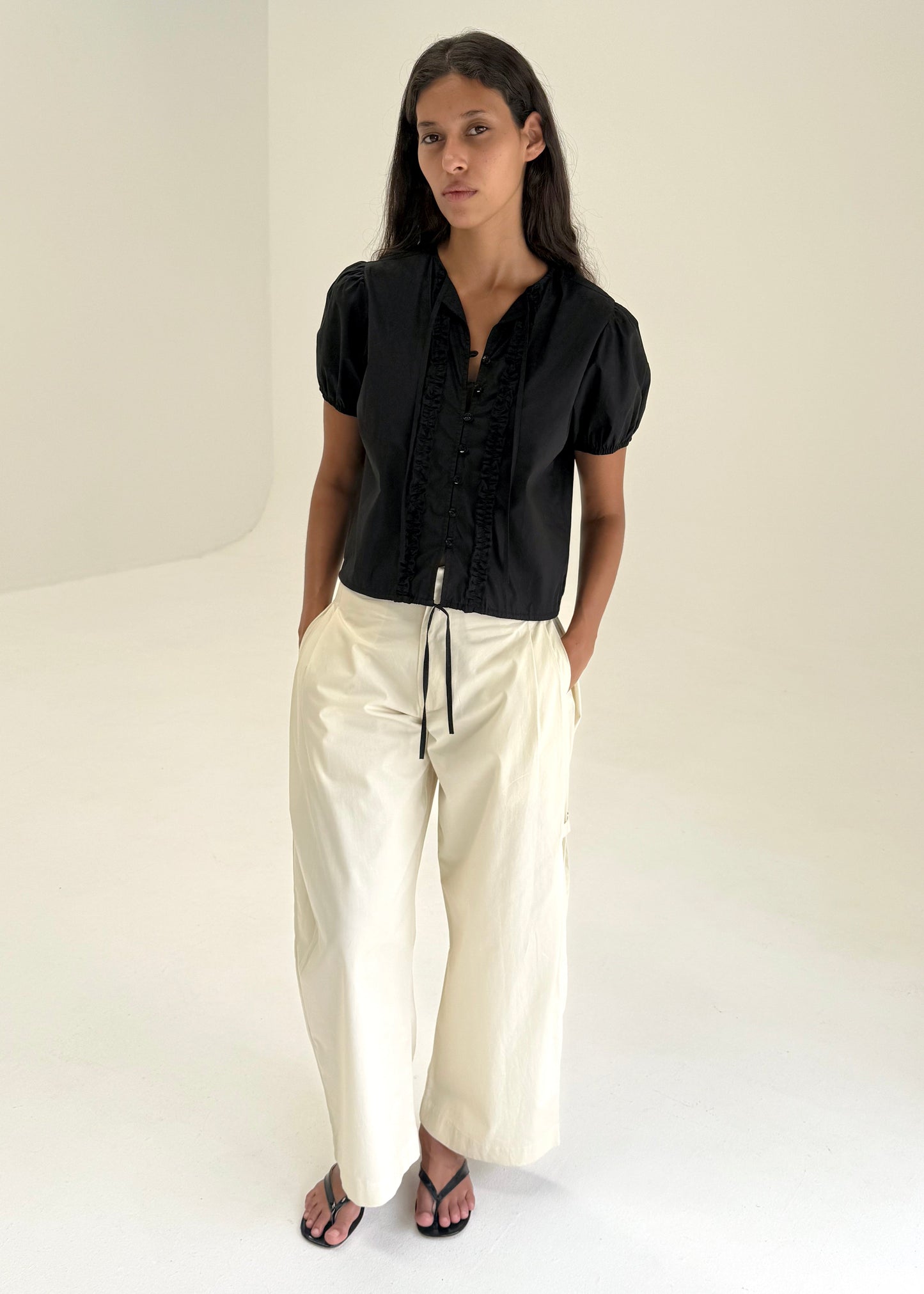 A woman with long dark hair stands in a minimalist, light-hued room wearing Deiji Studios' Double Frill Top - Black, loose white drawstring pants, and black sandals; her hands are casually tucked in her pockets.