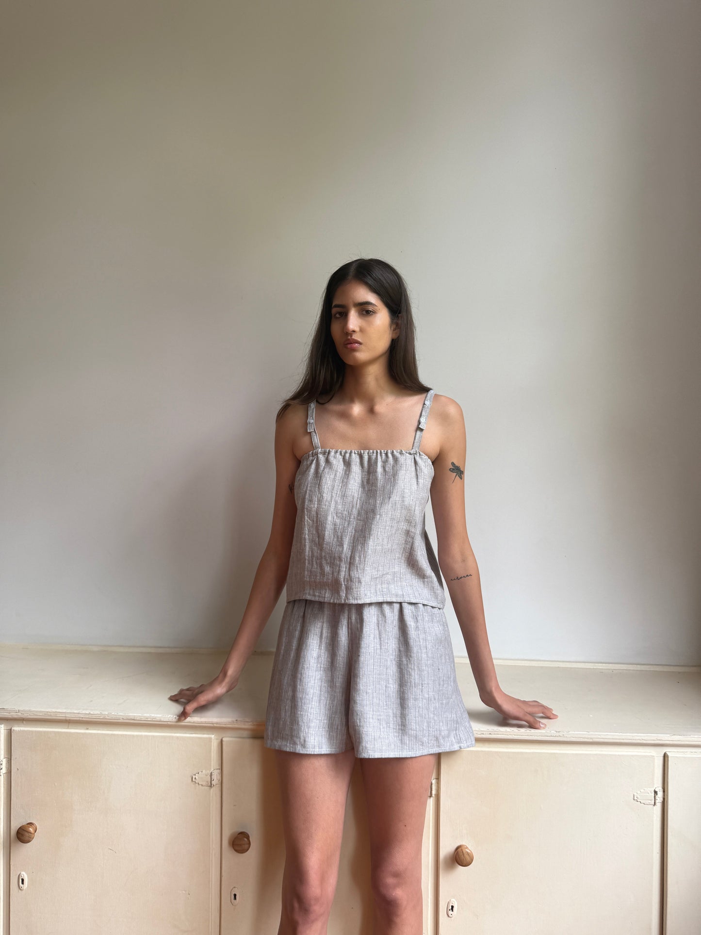 A woman with long dark hair stands against a light wall wearing the 0 Button Linen Set in Haze Blue, her hands resting on a beige cabinet as she gazes slightly to the side.
