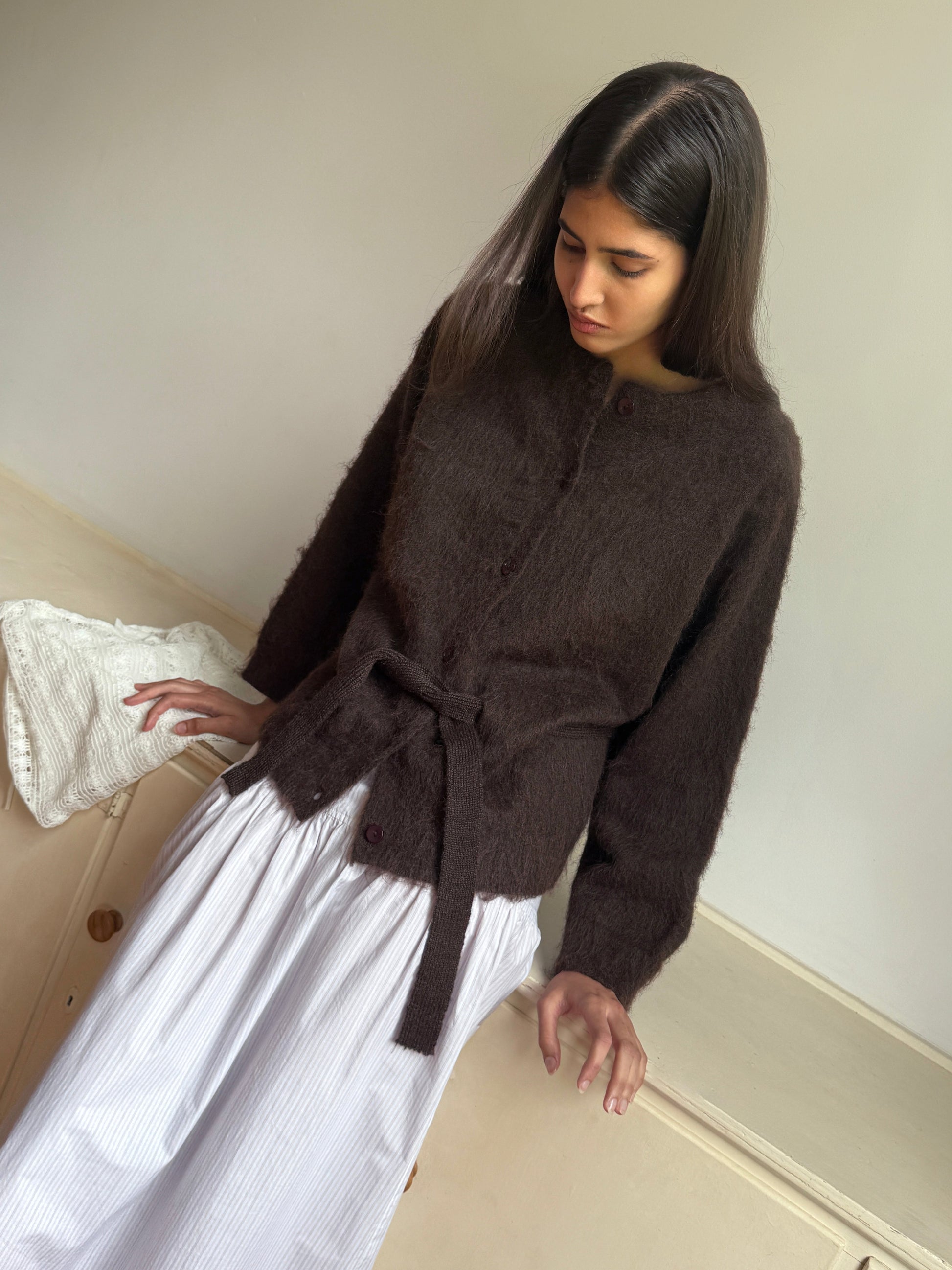 A woman with long dark hair wears the 0 Belted Cardi - Brown in an oversized fit, paired with a white pleated skirt, standing indoors and looking down calmly.