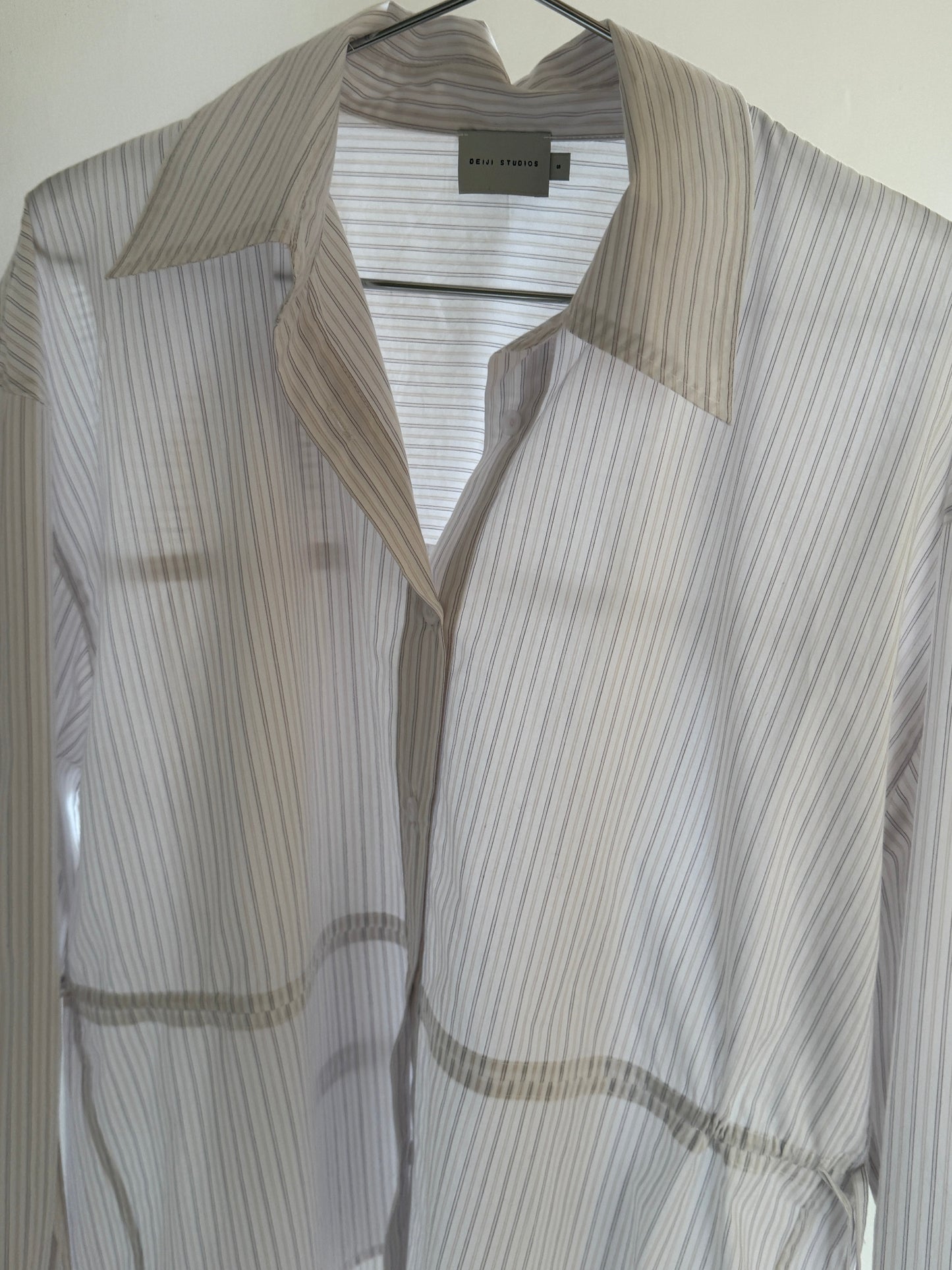 The Room Shirt - Pacific Stripe by 0 is a sheer, long-sleeved top made from organic cotton with thin light beige and white vertical stripes, shown hanging on a black hanger against a white background.