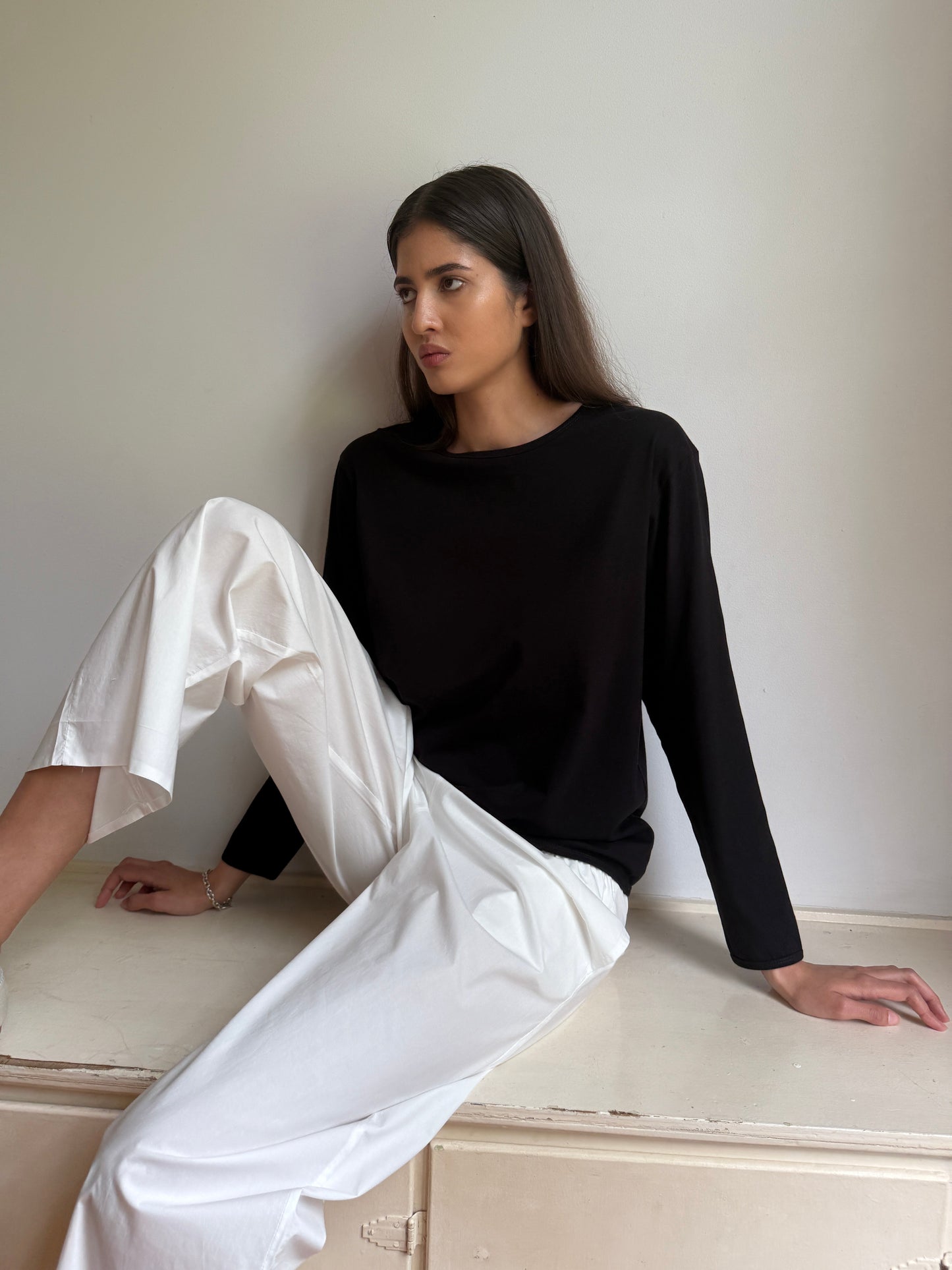 A woman with long dark hair sits against a light wall, wearing the Simple Long Sleeve - Black from 0 and white trousers, looking thoughtfully to the side.