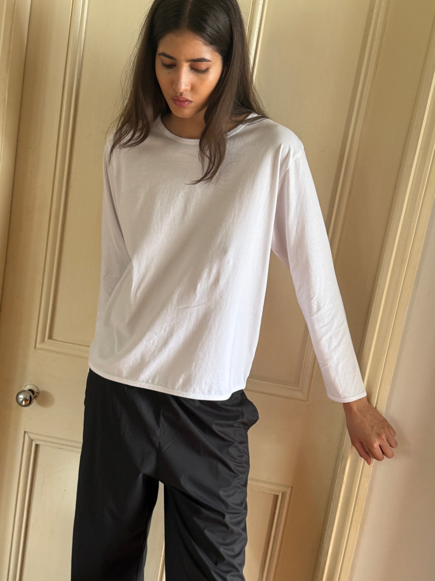A person with long, dark hair stands against a cream-colored door, wearing the Simple Long Sleeve - White by Deiji Studios and black pants, looking down with a neutral expression.