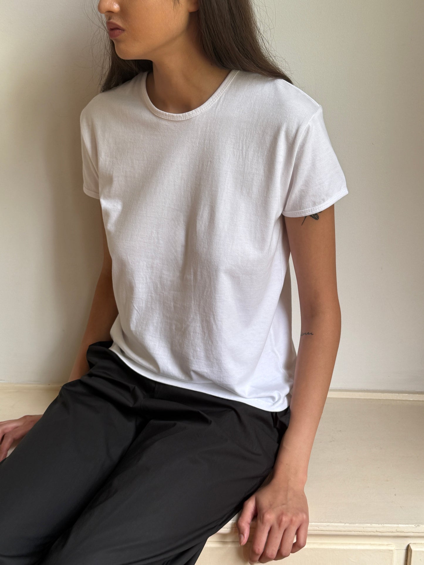 A person with long dark hair sits against a light-colored wall, wearing the Simple Tee - White in a relaxed fit and black pants. Their face is partially turned away, and their right arm rests on their leg.