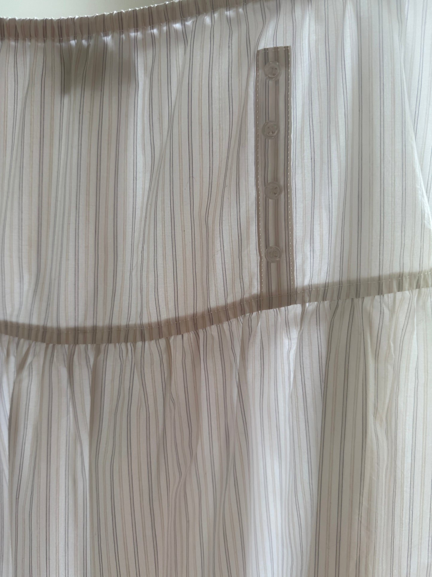 A close-up of the Button Skirt - Pacific Stripe shows sheer white fabric with thin beige stripes, a gathered seam, and a vertical row of four buttons on the placket—details suggesting an elegant, elastic-waist mid-length skirt.