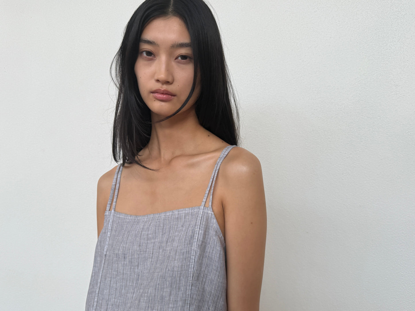 A woman with long black hair wears 0s sleeveless Linen Strap Dress in haze blue, featuring thin straps, as she stands against a plain white wall and calmly looks at the camera.