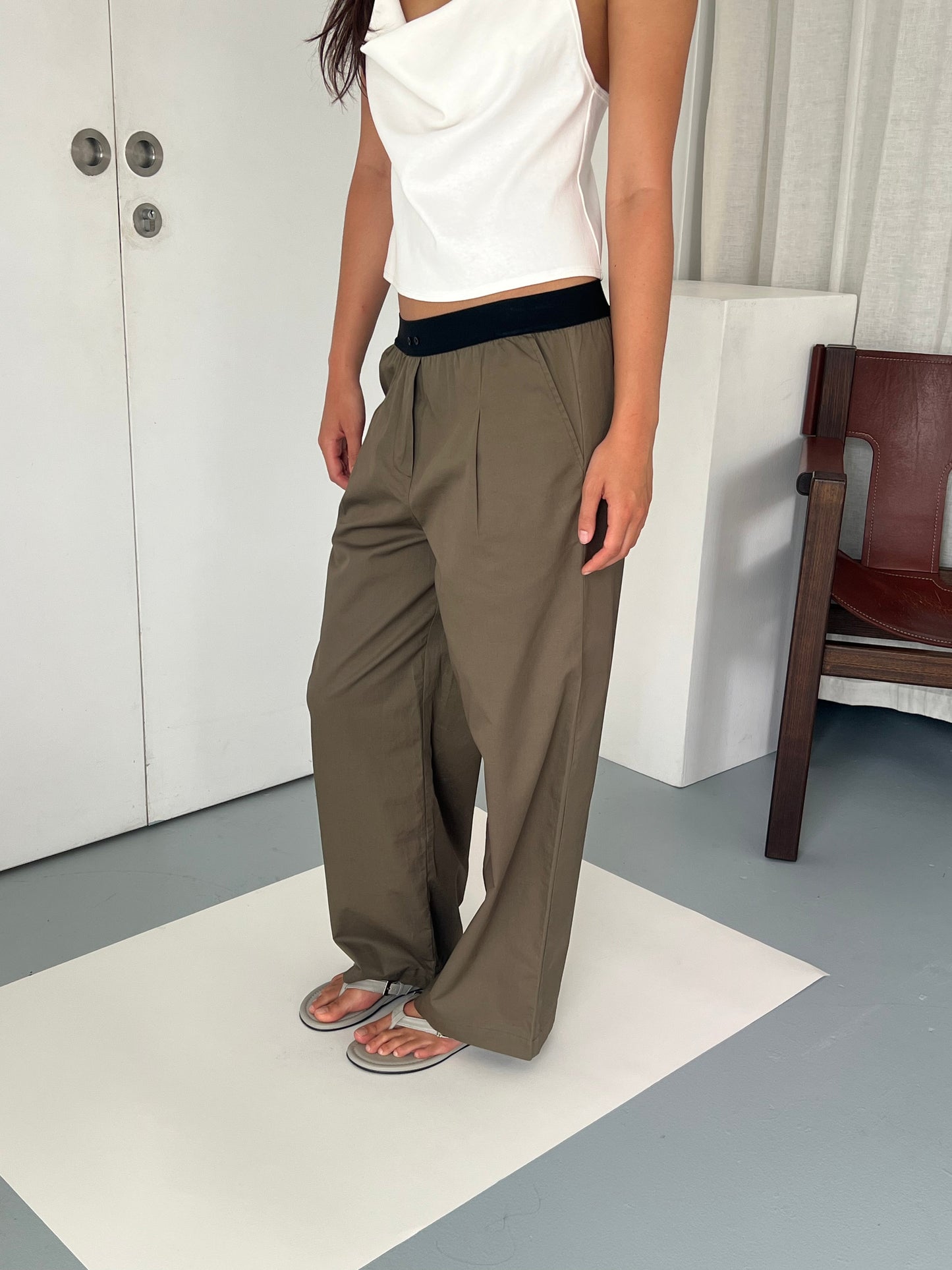 The Contrast Waist Pant - Olive by Deiji Studios features a relaxed straight leg and black contrast waist, styled on a female model with a white crop top and silver flip-flops.