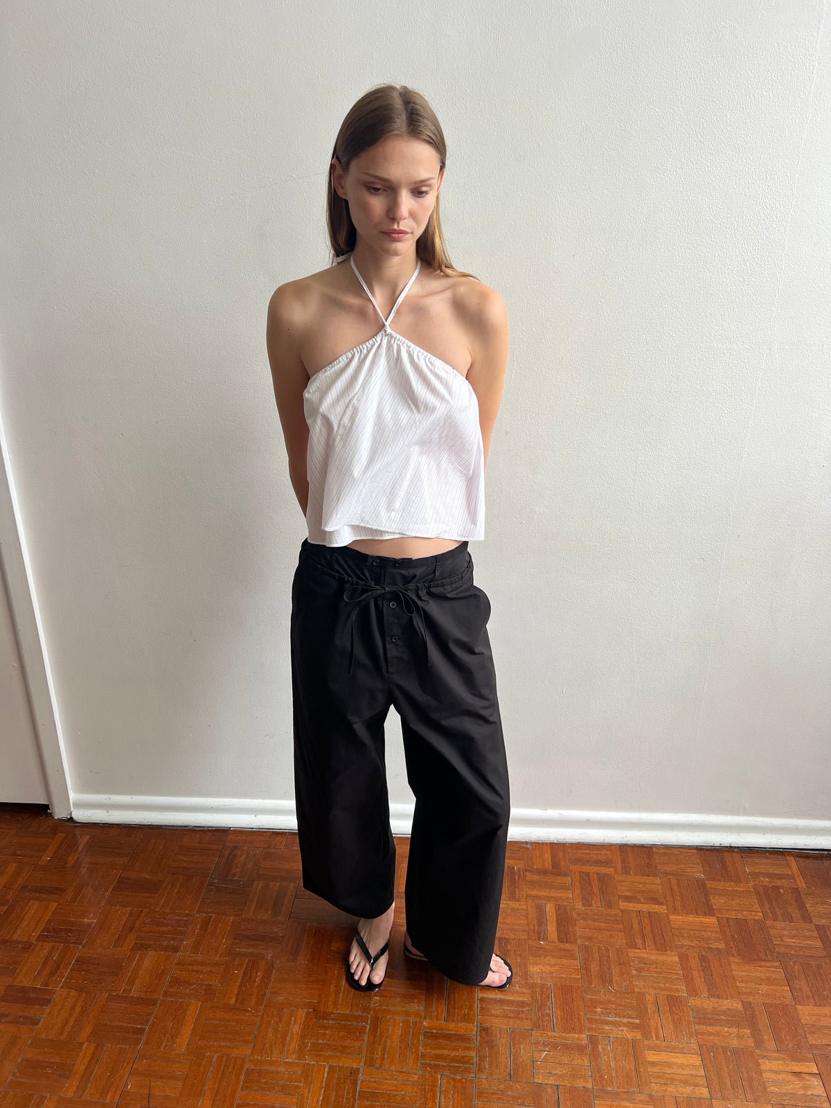 A woman stands indoors on a wooden floor, wearing a white halter top, Deiji Studios' Two Band Pant - Black, and black sandals. Her hands are behind her back as she looks down, with a plain white wall in the background.