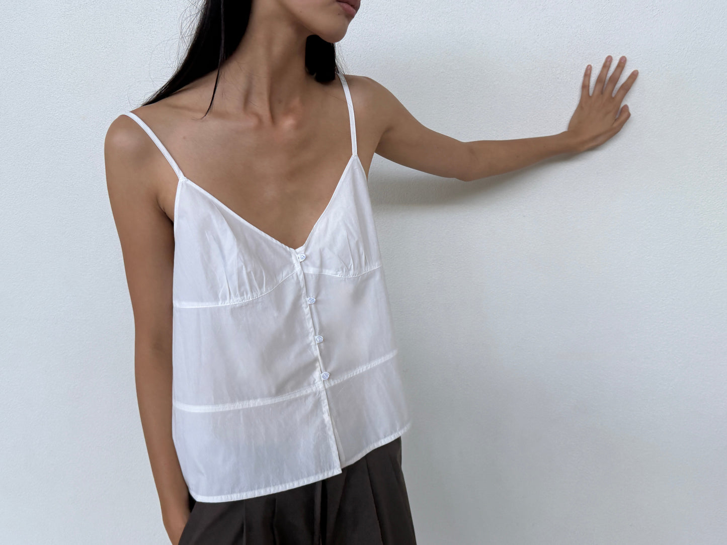 A woman in the white Seam Top by Deiji Studios and dark pants stands against a white wall with one hand resting on it, her long dark hair over her shoulder. The look reflects Deiji Studios’ signature minimalism.