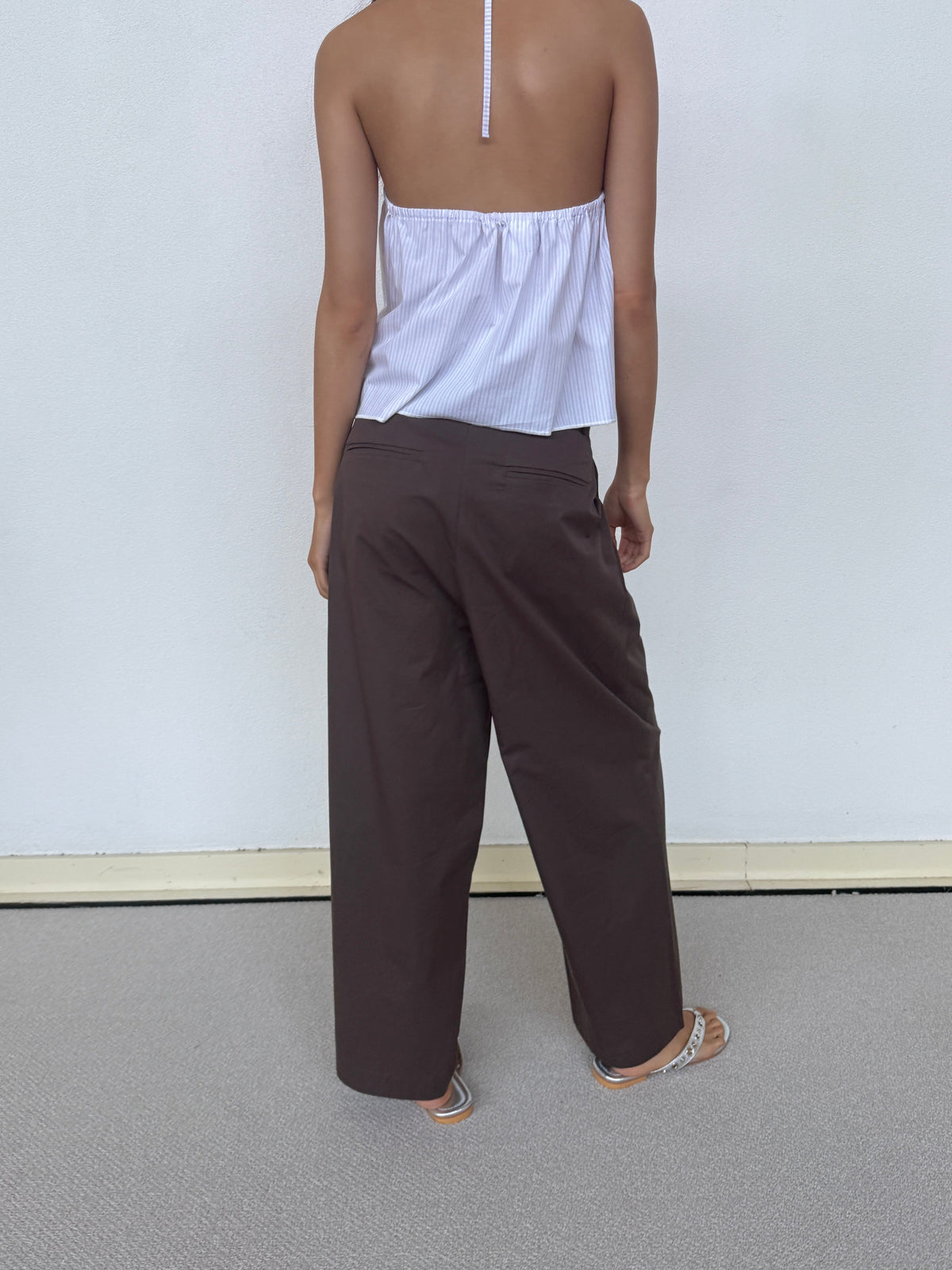 A person stands with their back to the camera, barefoot on a light carpet against a white wall, wearing Deiji Studios’ Turn Pant in Cocoa—relaxed and tailored with an adjustable drawcord—and a backless, halter-style white top.