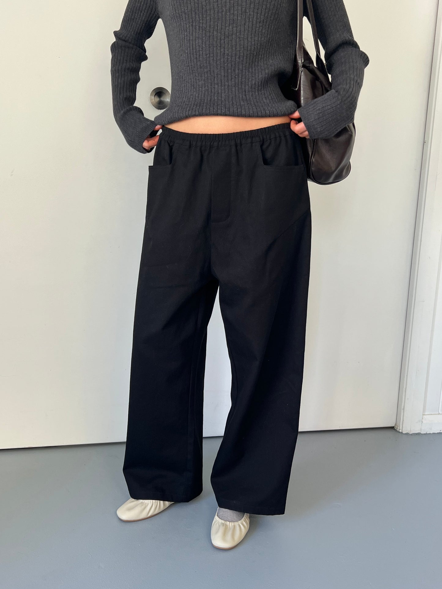 Wearing 0’s Square Pocket Pant in black, a person indoors pairs the wide-leg pants with a fitted gray ribbed sweater, cream flat shoes, and a black shoulder bag. The minimal setting includes a white door in the background.