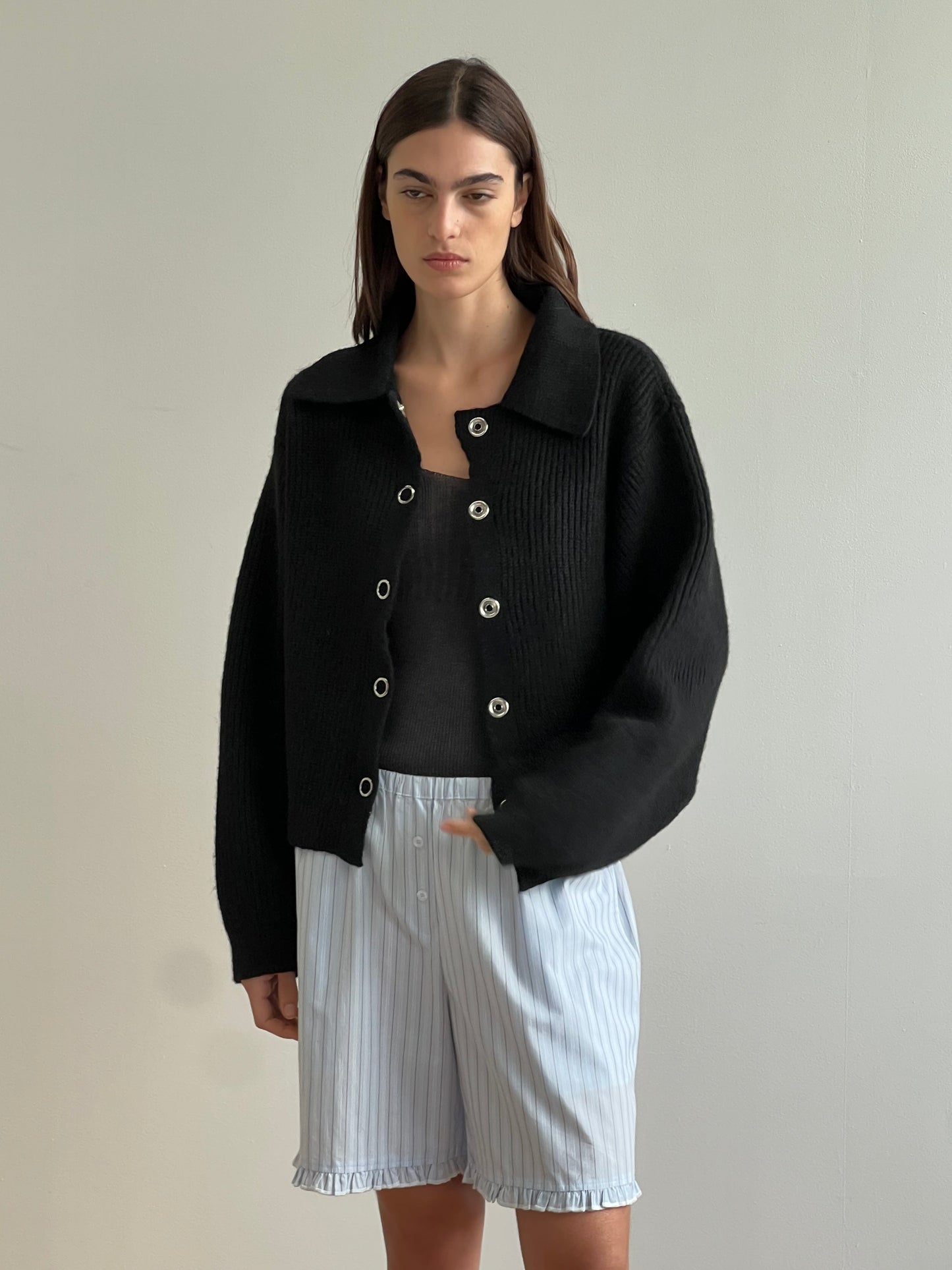 A woman with straight, dark hair and a neutral expression stands by a light wall, wearing the Stud Cardi Knit - Black over a black top and pinstriped, ruffled-hem shorts in light blue.