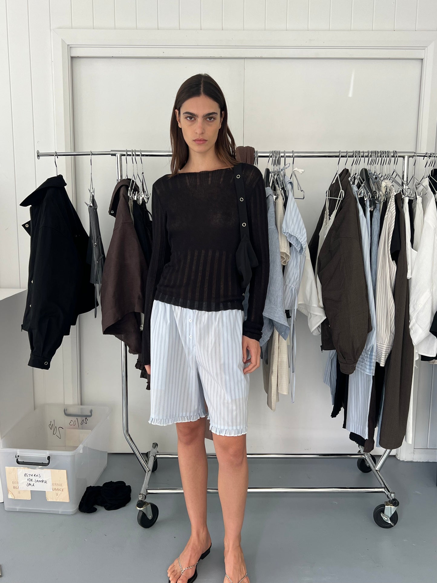 A woman with straight dark hair stands indoors by a clothing rack, barefoot, wearing the Open Work Longsleeve - Black and loose white pinstriped shorts. Clothes and a box are nearby.