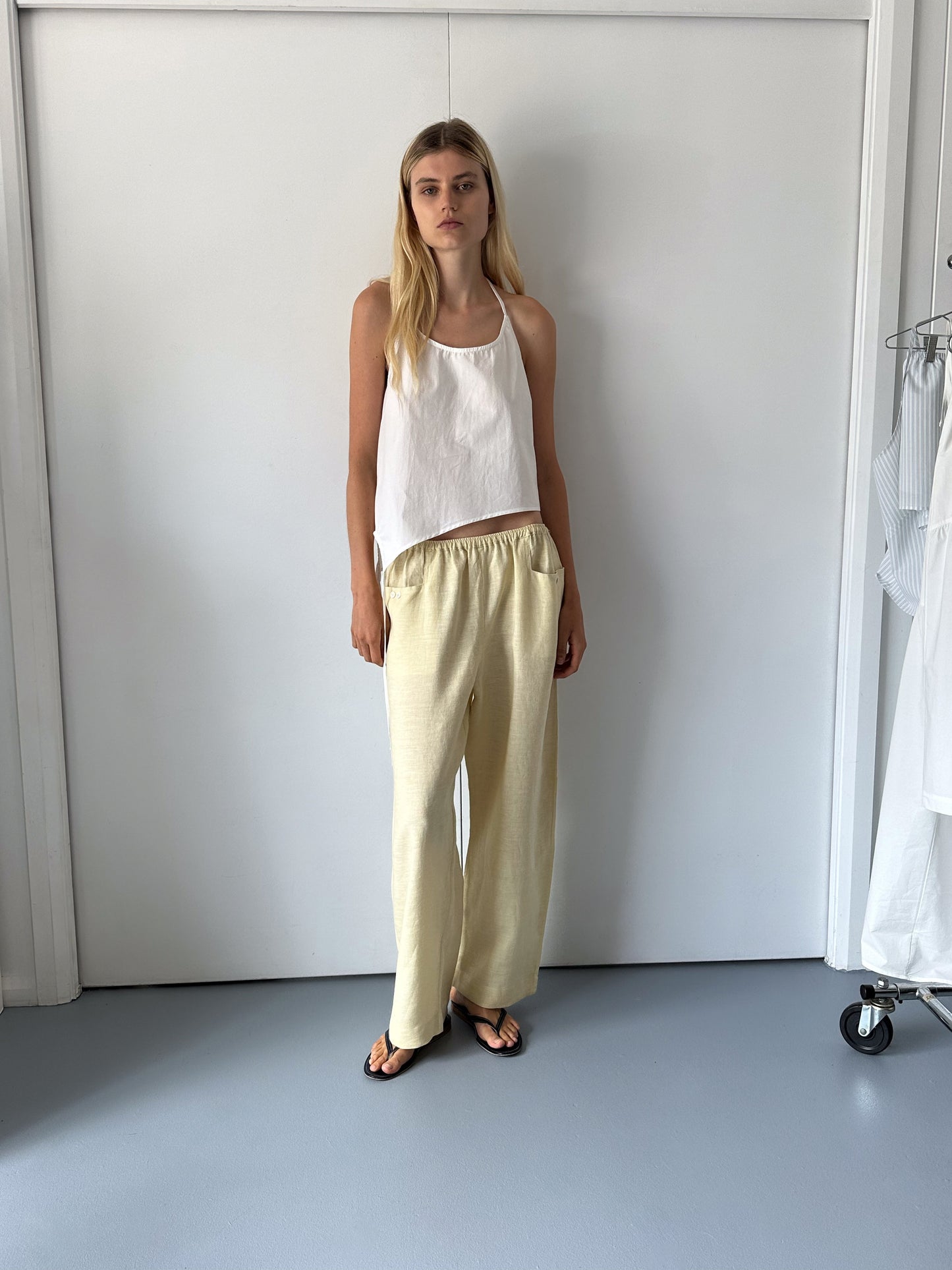 A woman with straight blonde hair stands indoors by a white wall wearing a white sleeveless top, the Linen Square Pocket Pant in Butter Yellow, and black sandals. To her right is a clothing rack with white garments.