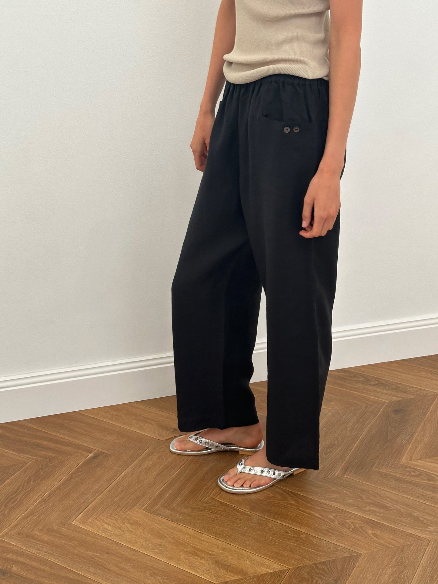 A person stands indoors on a wooden floor, wearing a beige sleeveless top and the Linen Square Pocket Pant - Black by Deiji Studios with an elastic waist and two buttons, paired with white sandals featuring silver studs. Their face is not visible.