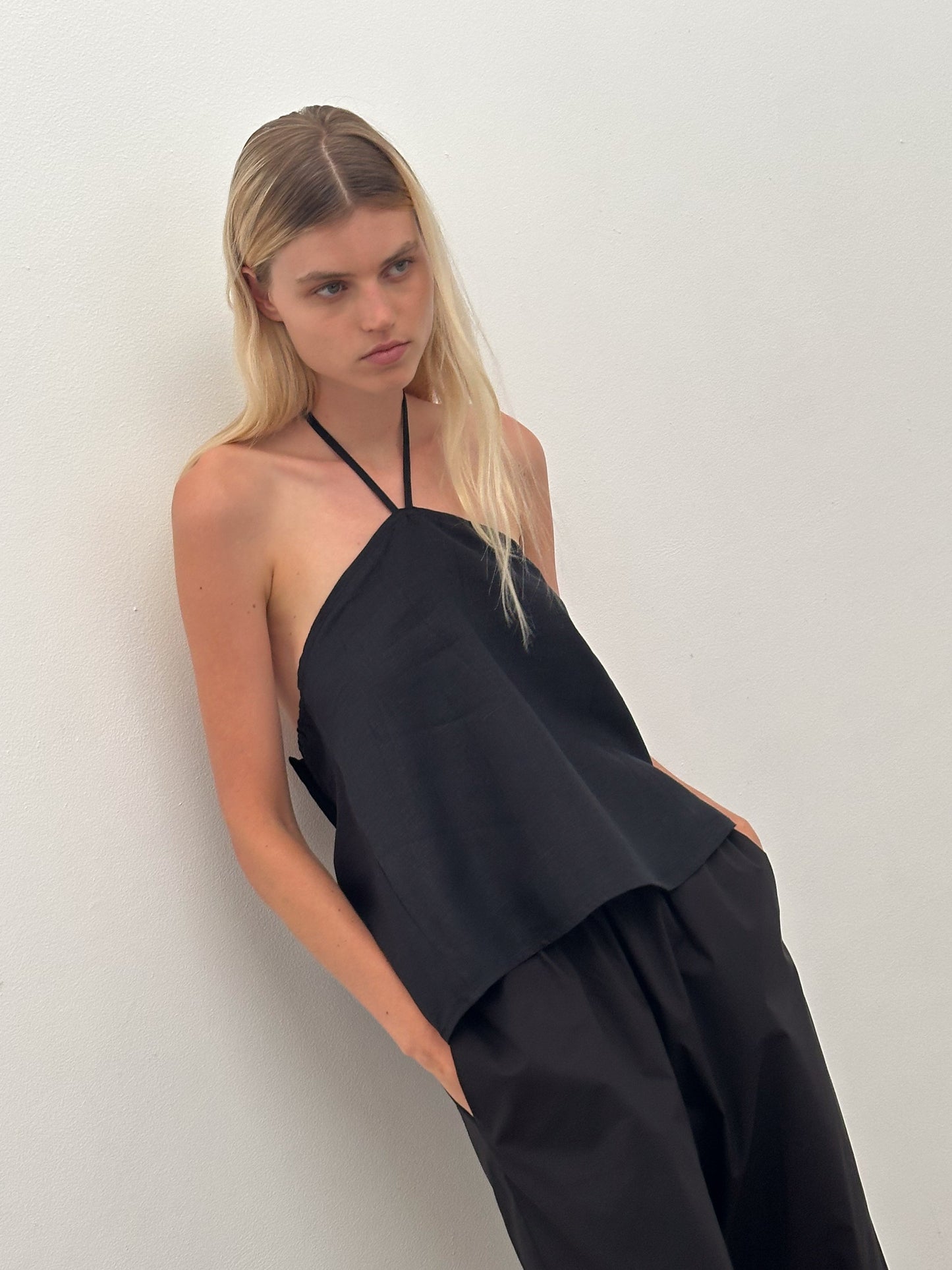 A young woman with long blonde hair wears a Deiji Studios Linen Wrap Back Top in black and loose black pants, standing against a plain white wall with her hands in her pockets, gazing downward with a neutral expression.
