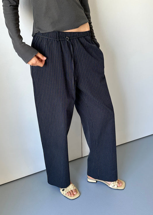 Someone stands against a light wall with hands in pockets, wearing the 0 Loop Pant - Ink Butter Stripe, a relaxed pinstripe trouser, paired with a dark gray long-sleeve top and cream heeled sandals. The head is not visible.