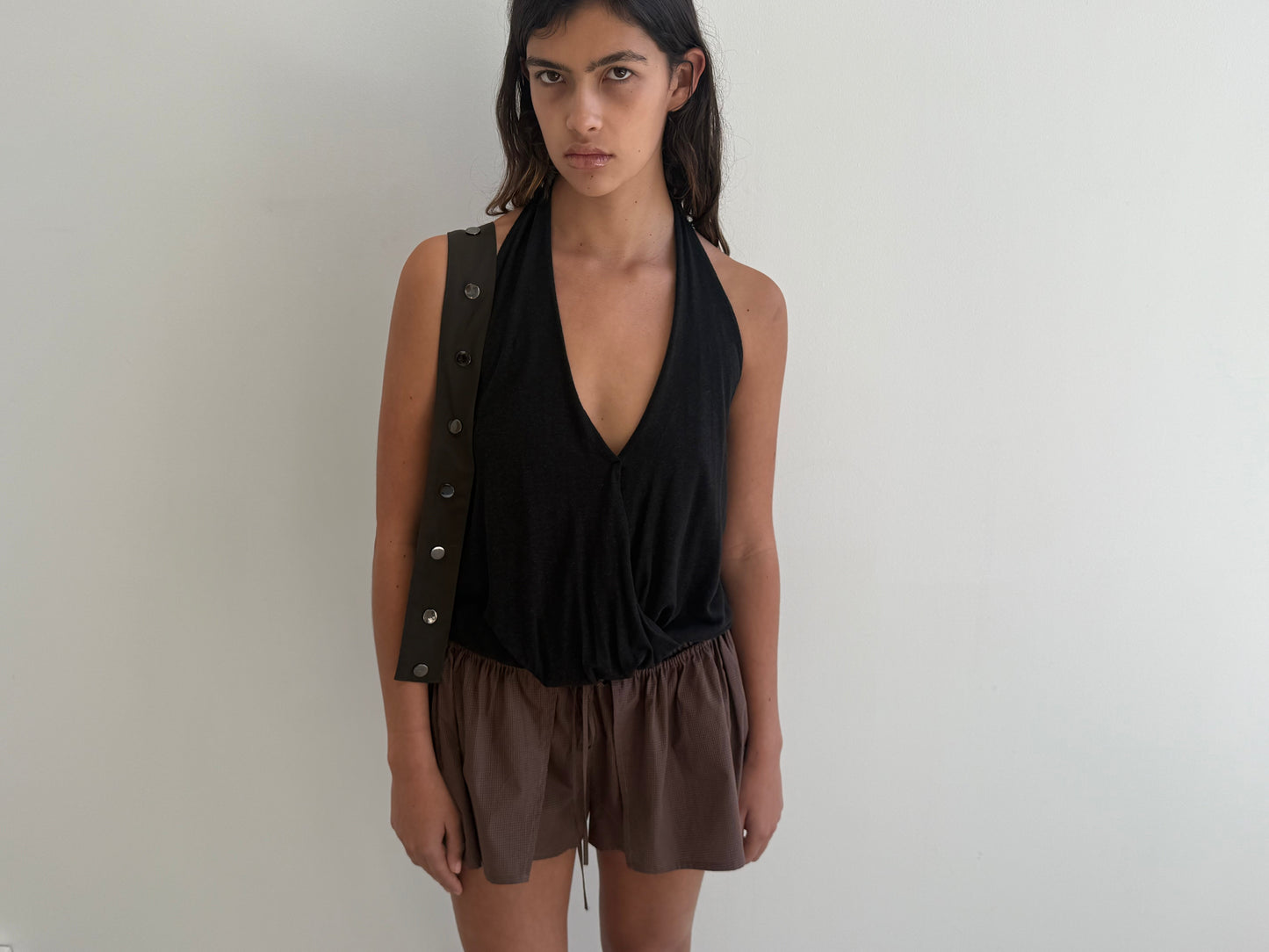 A person with long dark hair stands against a plain white wall, wearing a sleeveless black deep V-neck top and loose Carob Check skort by Deiji Studios, featuring a black strap with buttons over one shoulder.