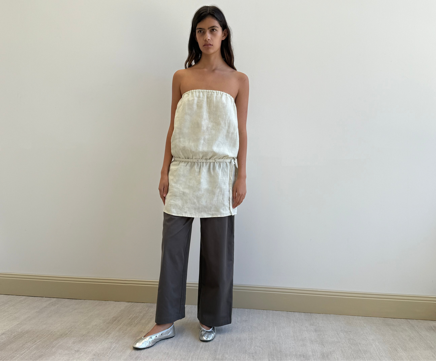 A woman with long dark hair and a neutral expression stands against a plain light wall, wearing the Strapless Linen Dress in Pastel Pistachio by Deiji Studios, cinched at the waist, paired with gray wide-leg pants and silver flats.