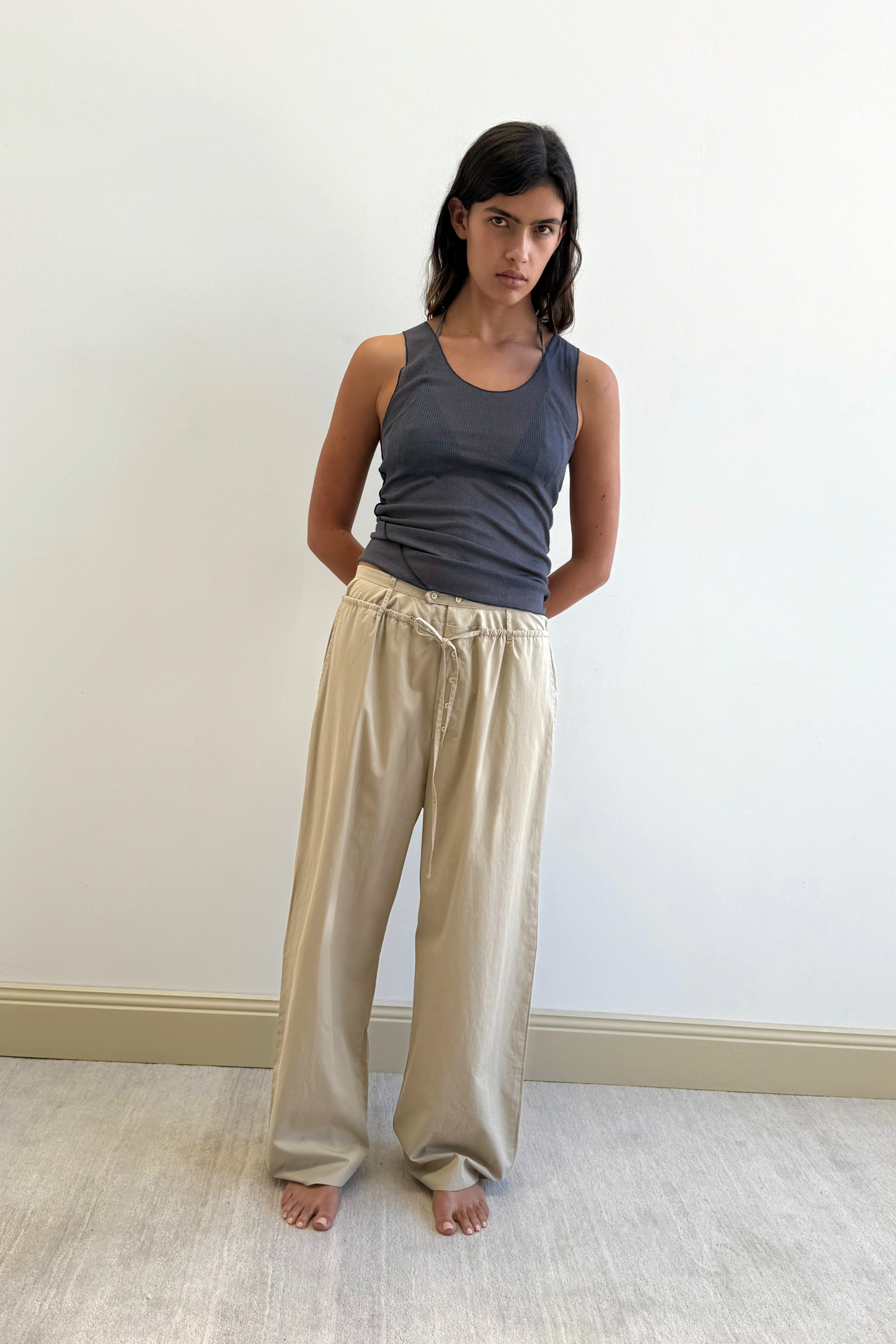A woman with long dark hair stands barefoot against a white wall, wearing a sleeveless gray top and the Two Band Pant - Fawn by Deiji Studios. Her arms are behind her back and her expression is neutral.