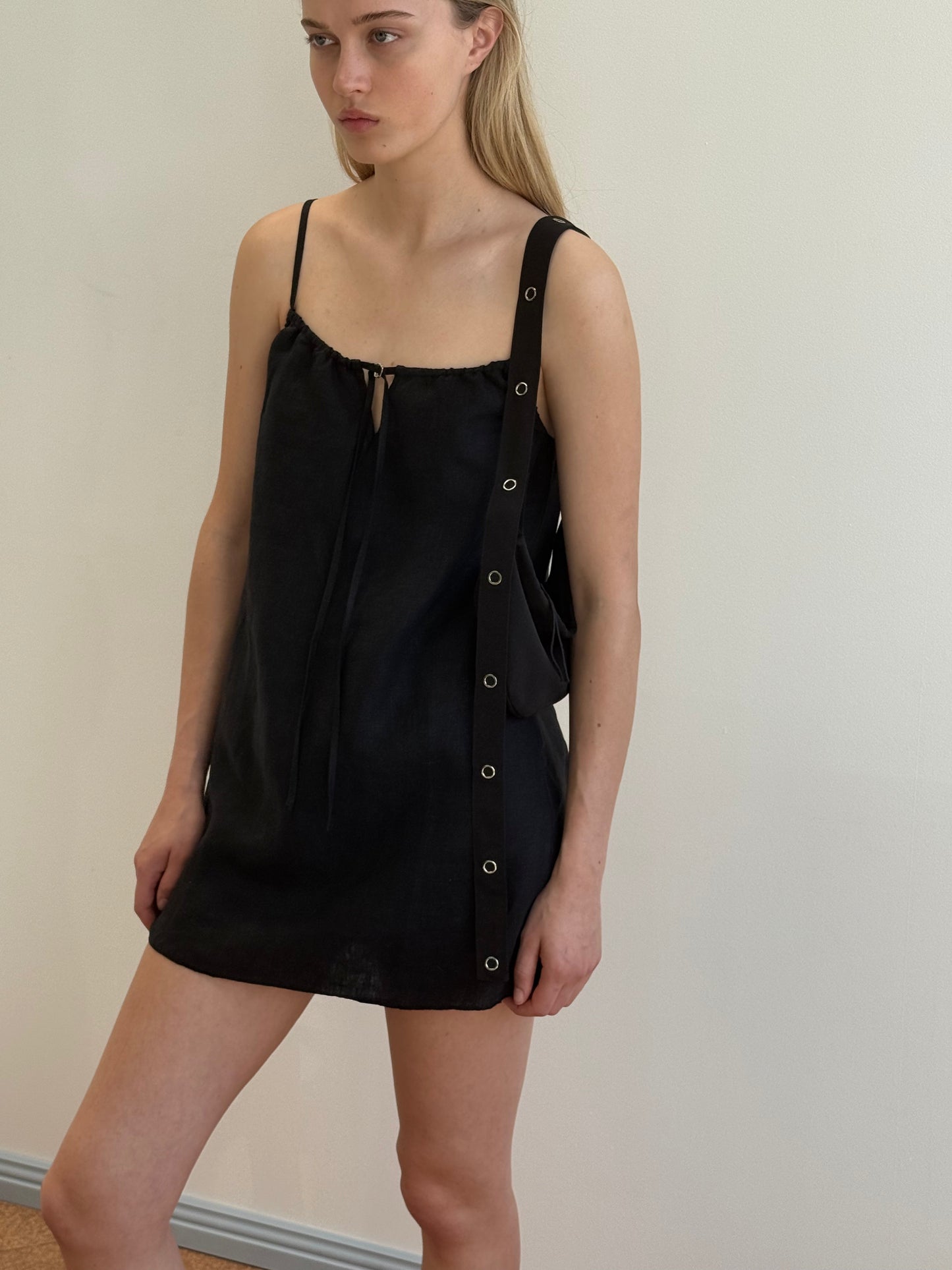 A woman with straight light brown hair and a neutral expression stands against a plain light wall, modeling the Lettuce Mini Dress - Black with thin straps, grommet details, and tie neckline—a look inspired by Deiji Studios’ modern minimalism.
