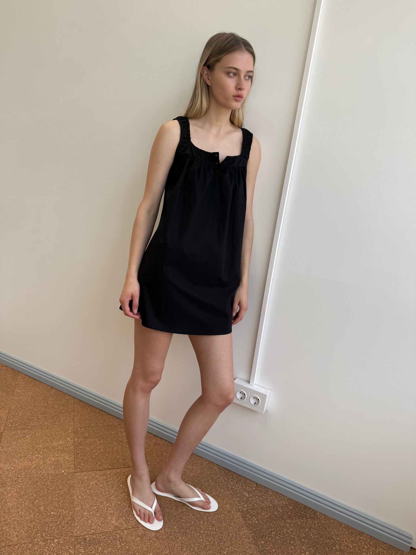 A woman stands indoors against a white wall, wearing the Yoke Mini Dress - Black by Deiji Studios and white flip-flops. She has straight light brown hair, a neutral expression, and her arms are relaxed at her sides.