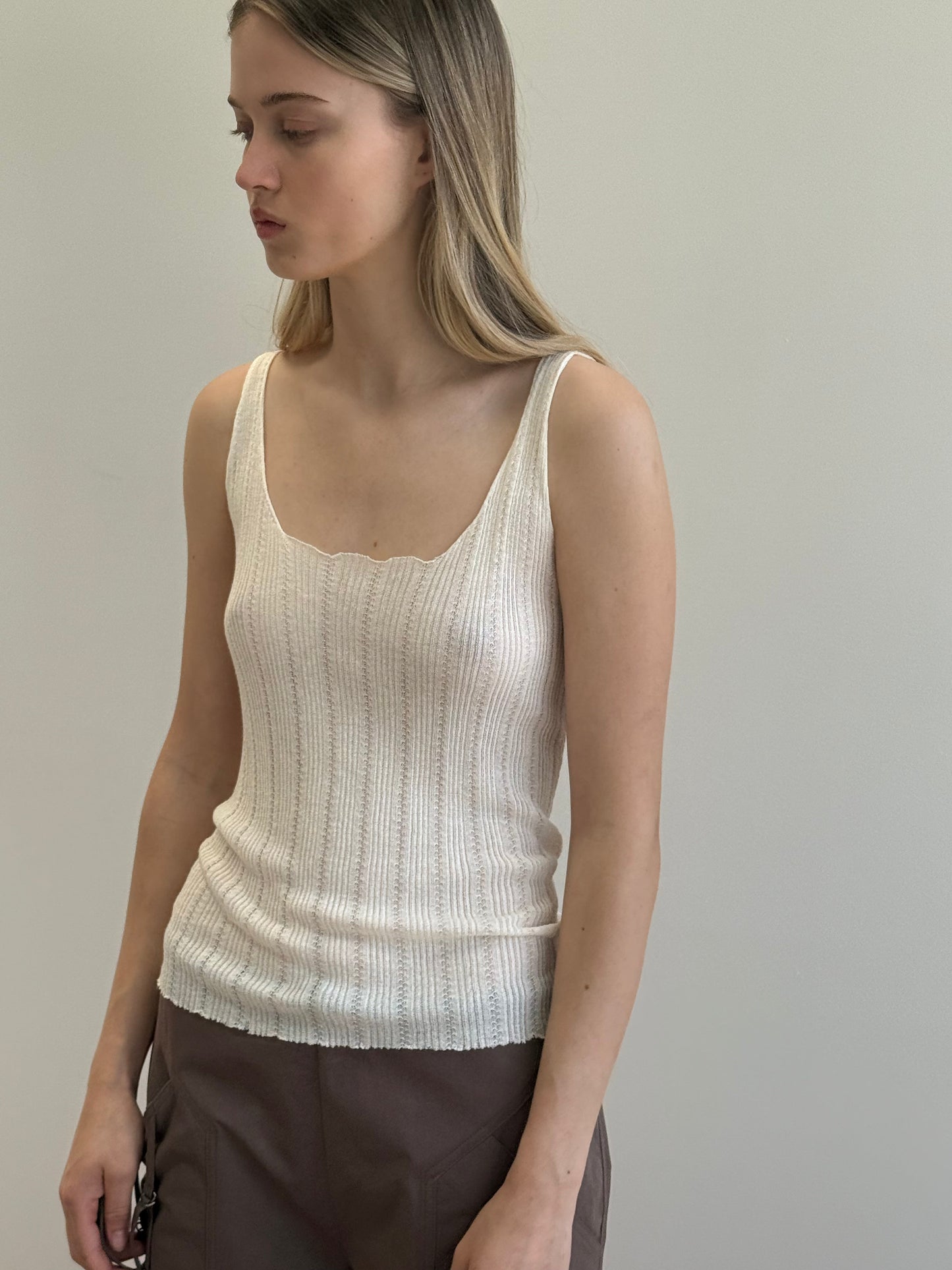 A woman with long, straight hair wears the Open Work Tank - White from Deiji Studios and brown pants, standing against a plain light background, looking slightly down and to the side.