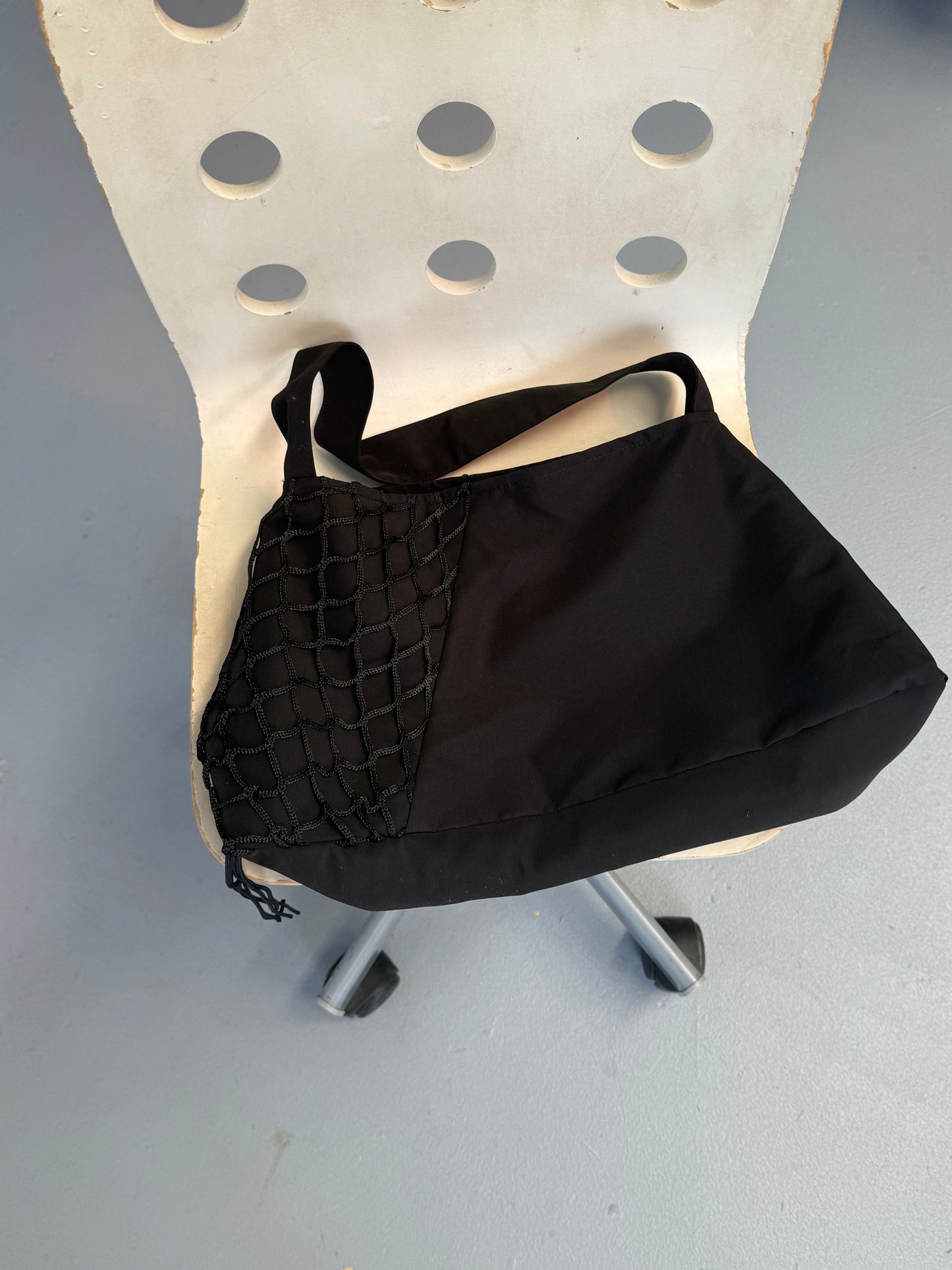 The Net Contrast Bag - Black, featuring a netted pocket, sits on a white chair with circular cutouts in the backrest on a light gray floor.
