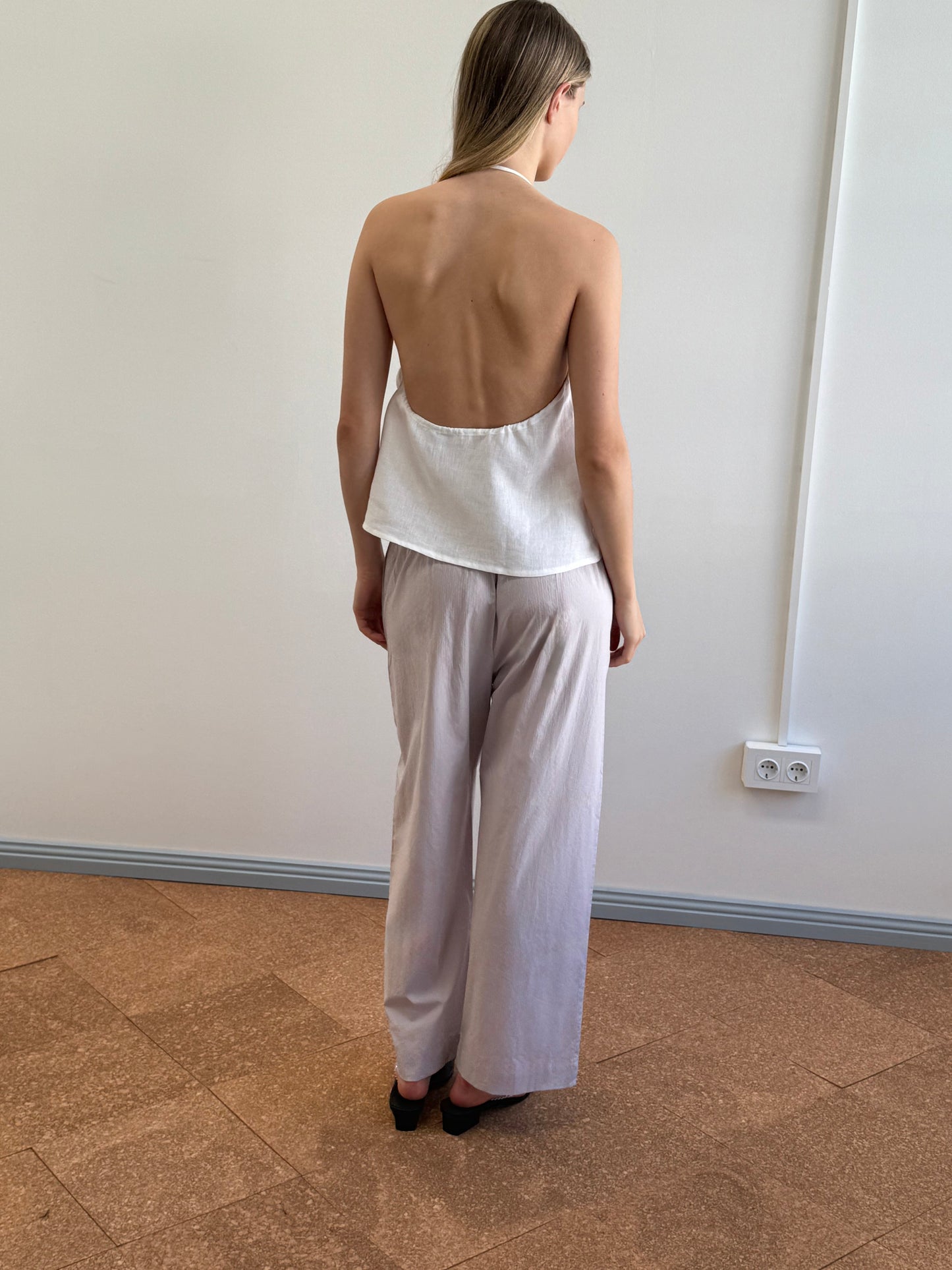 A woman with straight, shoulder-length hair faces away from the camera indoors, wearing the O Ring Halter - White top and loose white pants, paired with black shoes. A plain wall and electrical outlet appear in the background.