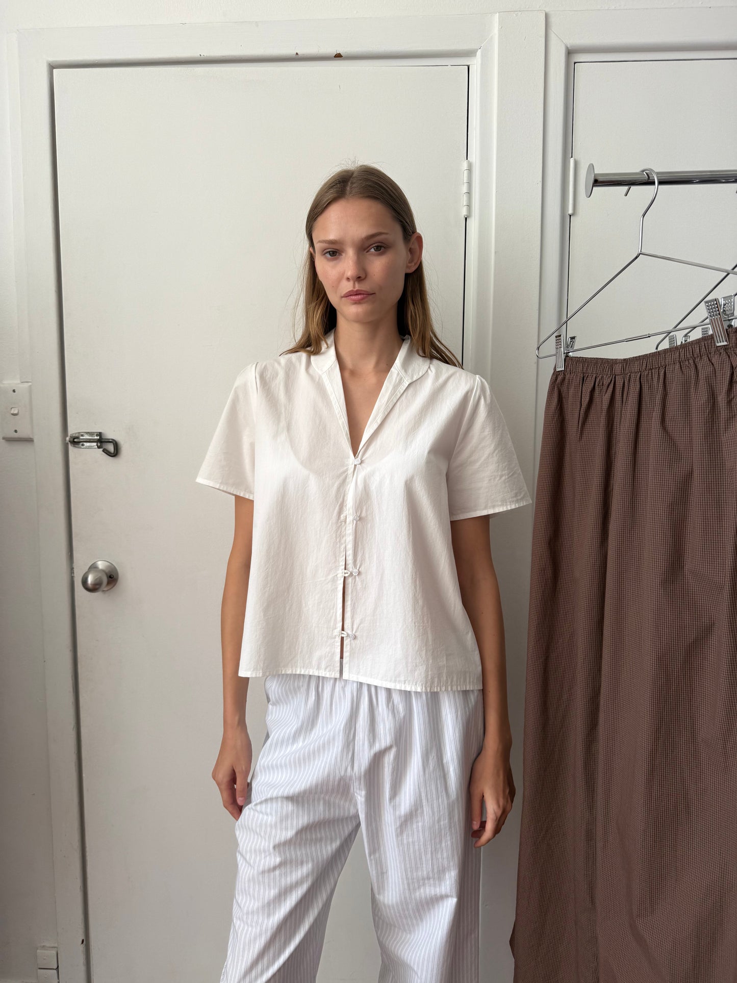A woman stands indoors wearing the Sails Top - White with white striped pants. A brown skirt hangs on a nearby rack, and a white door and wall are in the background.