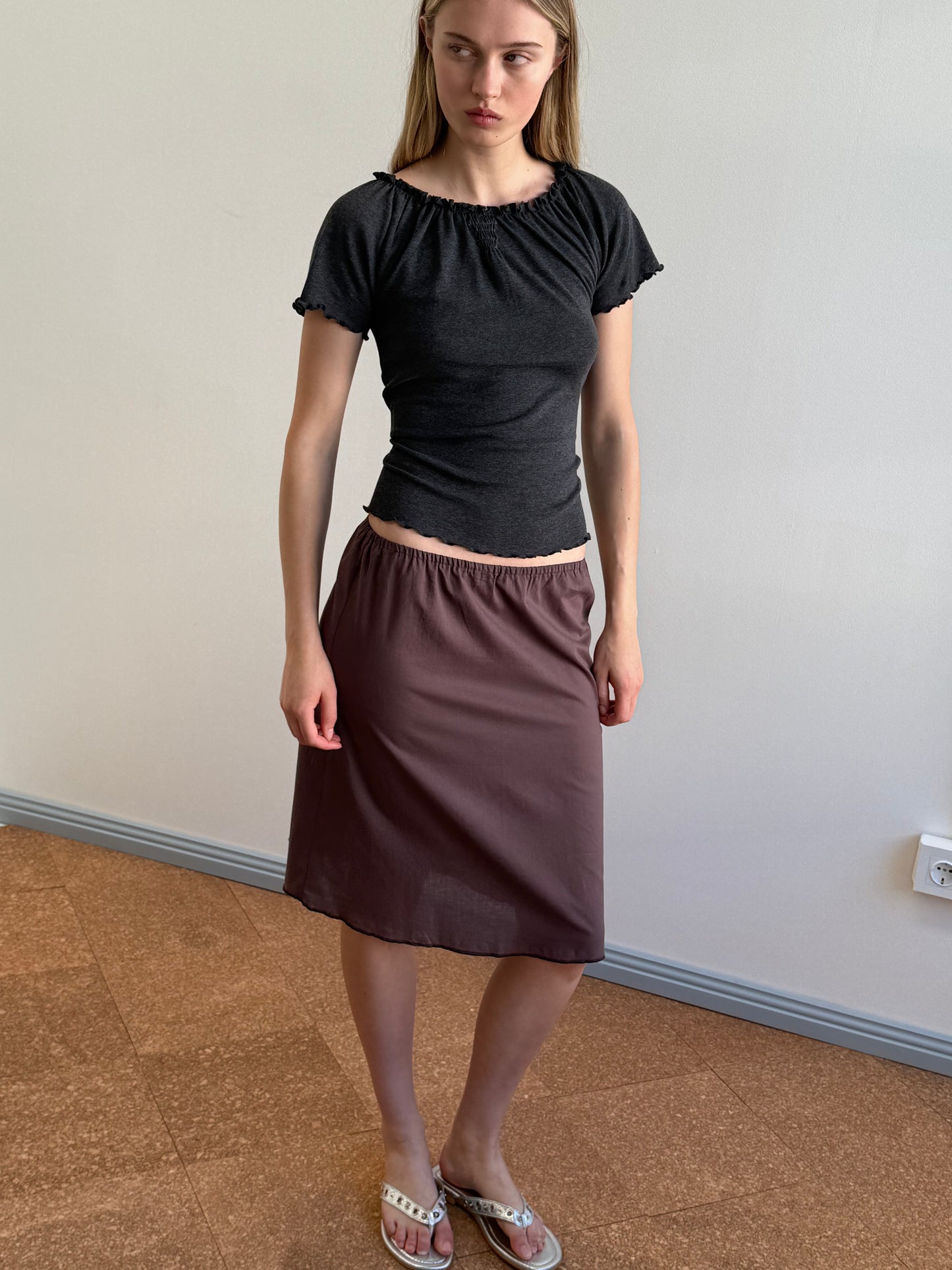 A woman with long blonde hair stands indoors wearing sandals, a fitted black t-shirt, and the Simple Skirt in Raisin, a knee-length everyday skirt. She looks slightly to the side with a neutral expression.