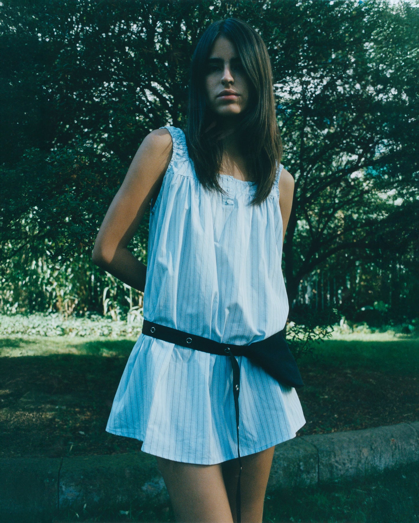 A young woman with long brown hair and a neutral expression stands outdoors before leafy trees, wearing a loose, sleeveless, light blue dress with subtle stripes and the Snap Belt Bag - Black by her waist.
