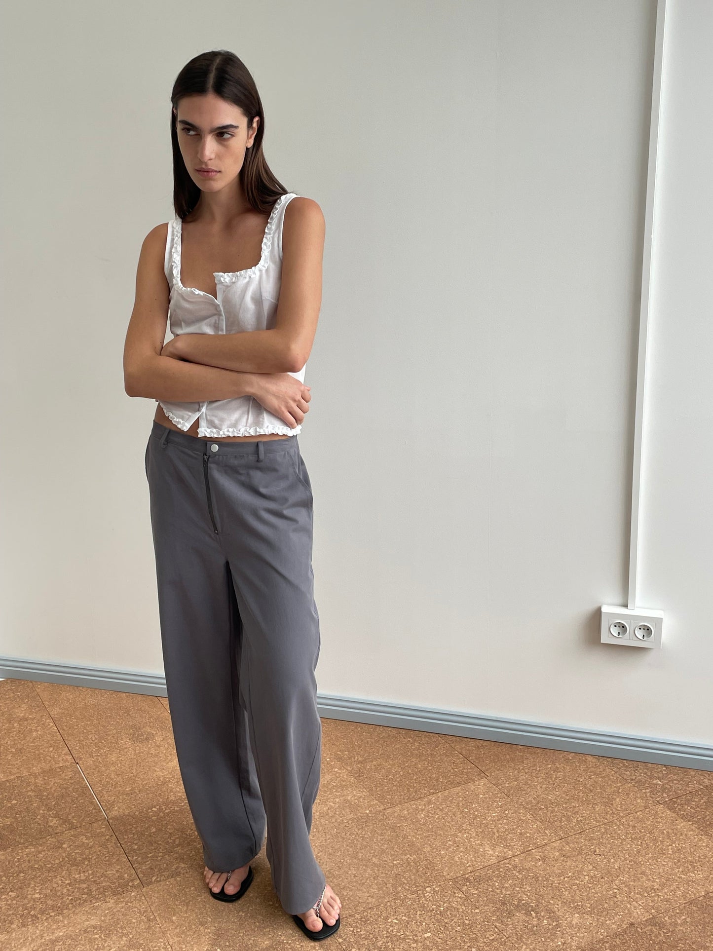 A woman stands indoors against a plain wall, wearing the Deiji Studios Button Frill Top in white with lace trim, gray wide-leg pants, and black sandals. Her arms are crossed, and she has a serious expression.