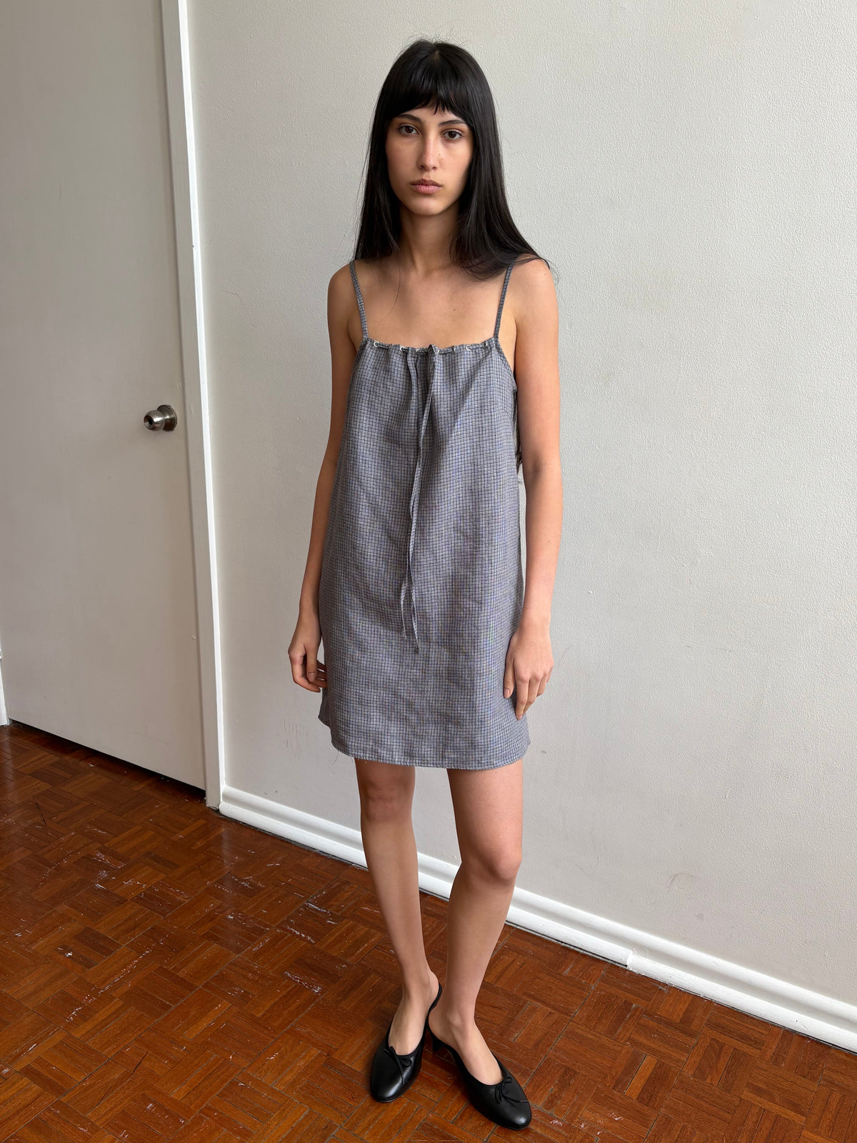 A woman with dark hair and a fringe stands in a room wearing the Weave Mini - Indigo Check by Deiji Studios, an indigo micro-check linen mini dress with thin straps and a drawstring front tie, with black flats.