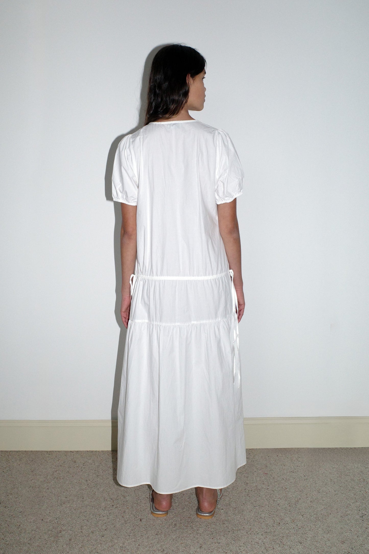 A woman stands with her back to the camera on a beige carpet, wearing the Waistcord Dress - White by Deiji Studios—an ankle-length, short-sleeved organic cotton dress—and beige heels against a white wall.