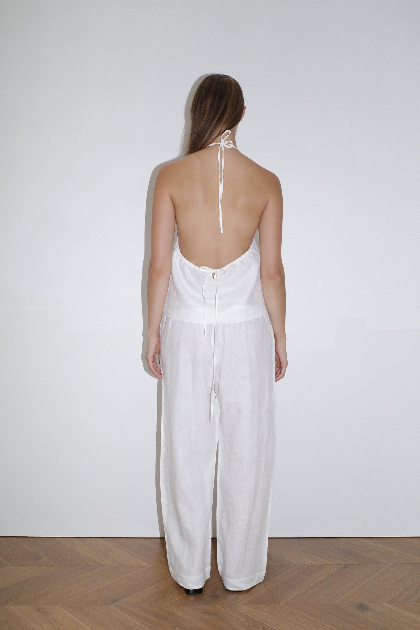 A person stands on a wooden floor facing a white wall, wearing a white halter-neck, backless top and the Linen Square Pocket Pant - White by Deiji Studios in organic French linen. Their straight hair is worn down.